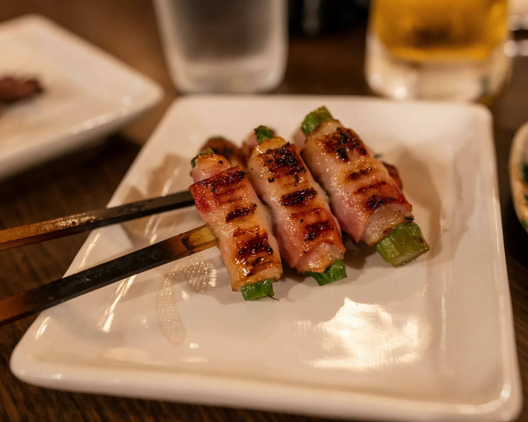 Three asparagus and bacon skewers, striped from the grill, lifted from a white plate with chopsticks