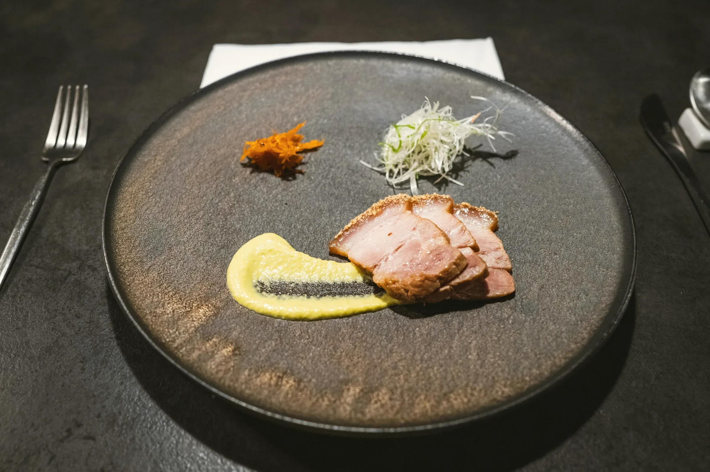 Slices of smoked duck with kabocha purée, shredded leek and a chili relish on a dark stoneware plate