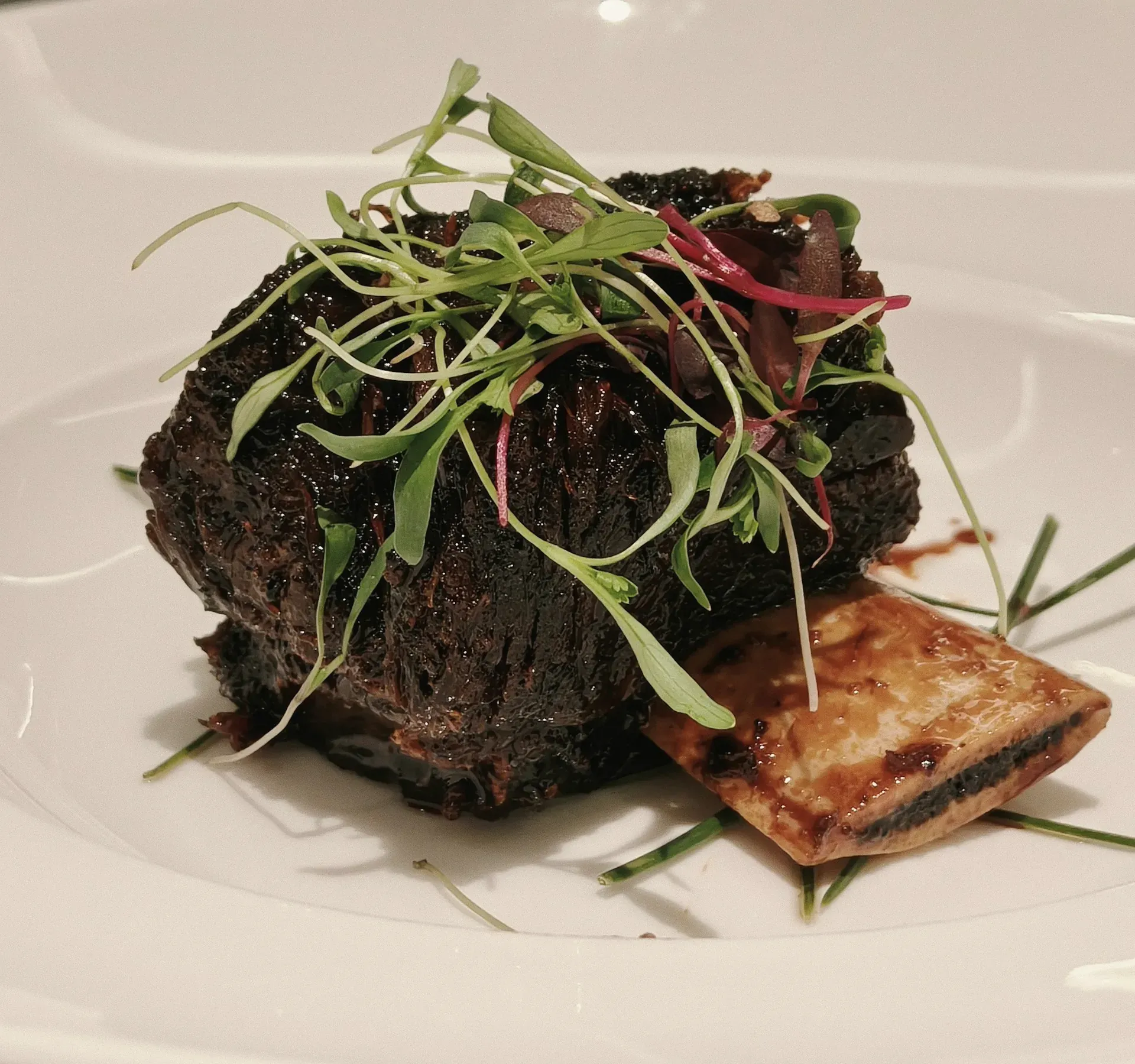 A braised short rib under microgreens, its bone beside it, in a wide white bowl