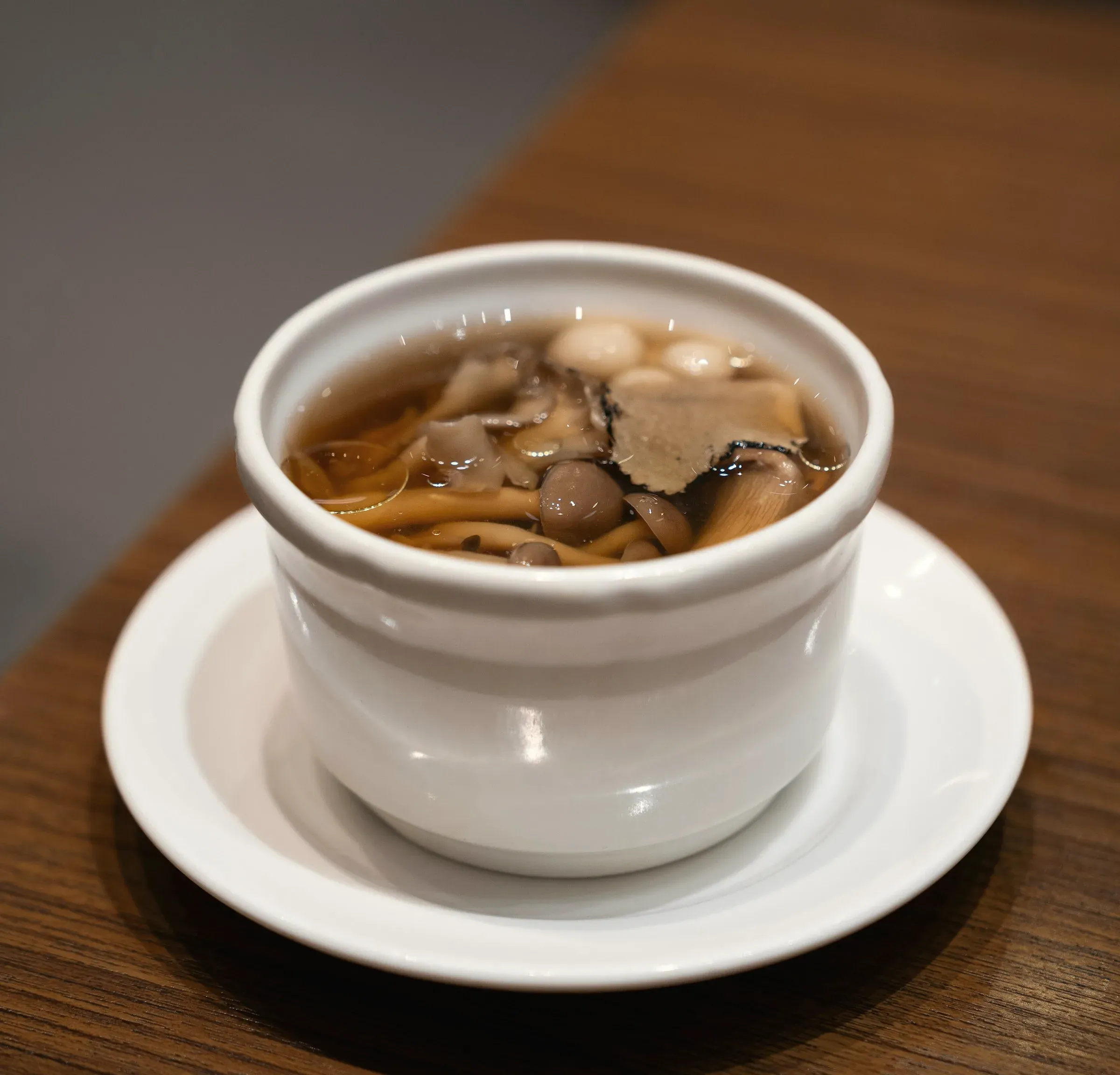 Shimeji and oyster mushrooms in a clear amber broth under a shaving of black truffle, in a white porcelain cup