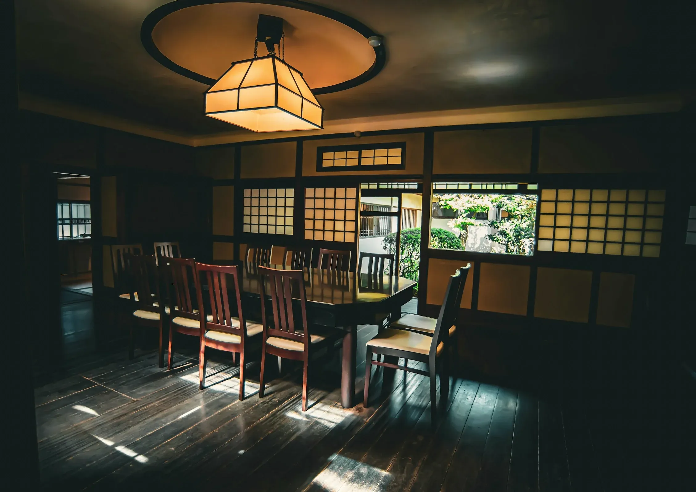 A long dark table set with twelve chairs under a paper lantern, shoji windows open onto the courtyard garden