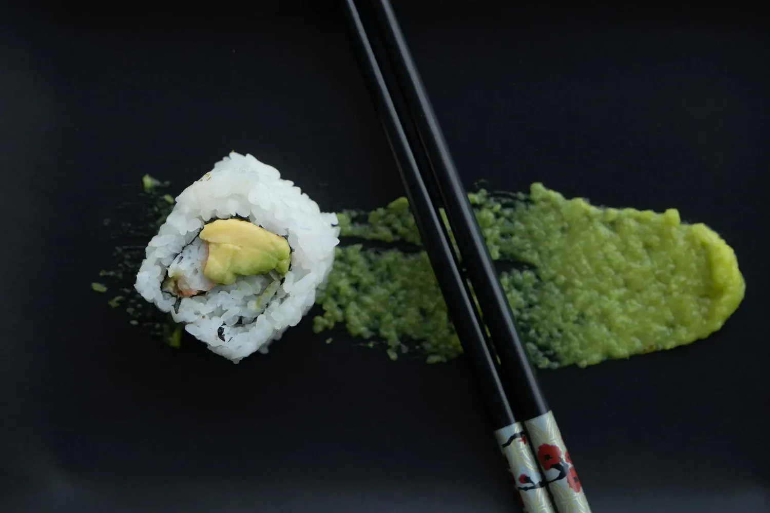One piece of California roll with crab and avocado on a black plate beside chopsticks and wasabi