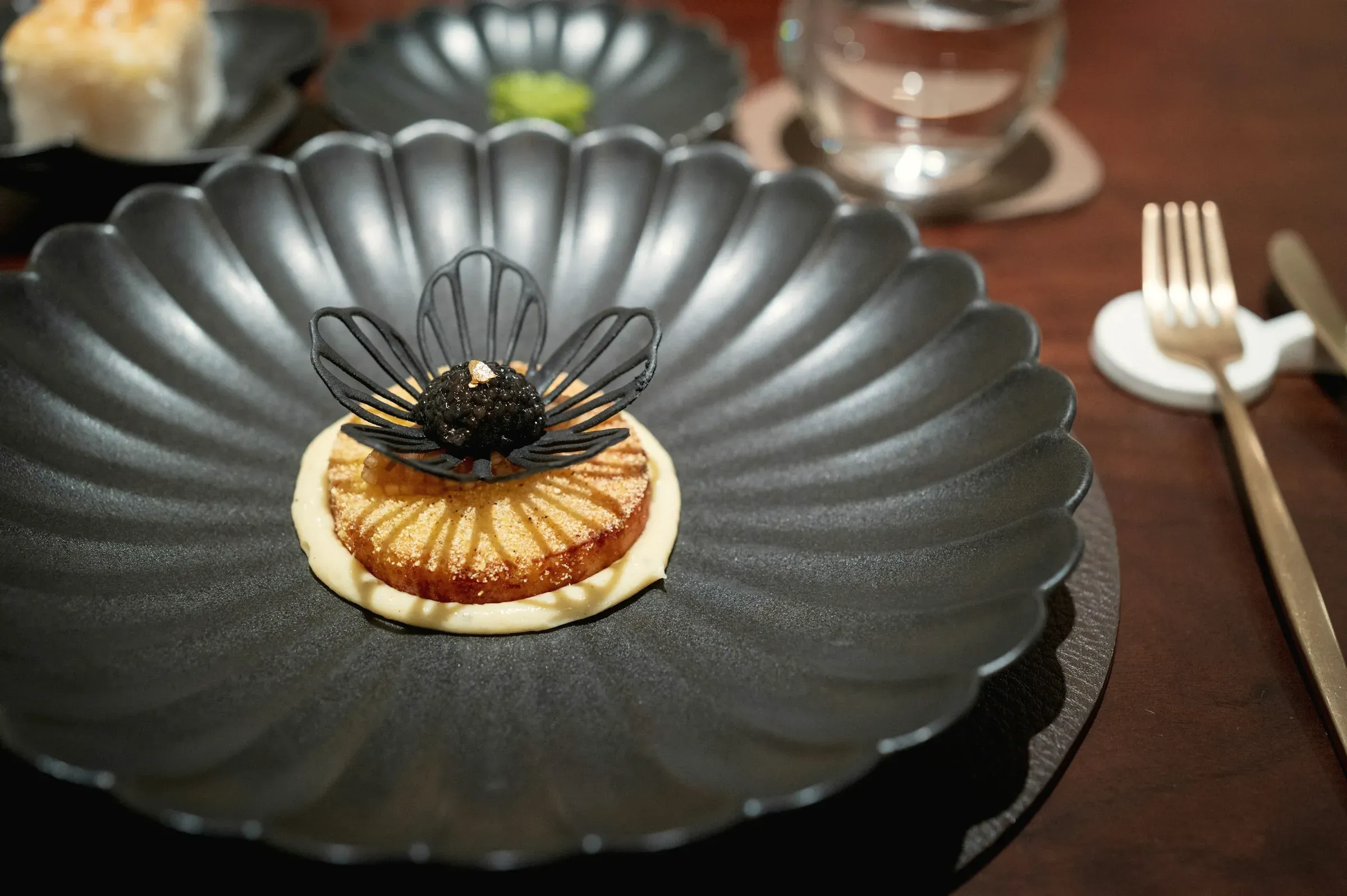 Caviar on a golden round cake under a black flower-shaped tuile, on a dark scalloped plate