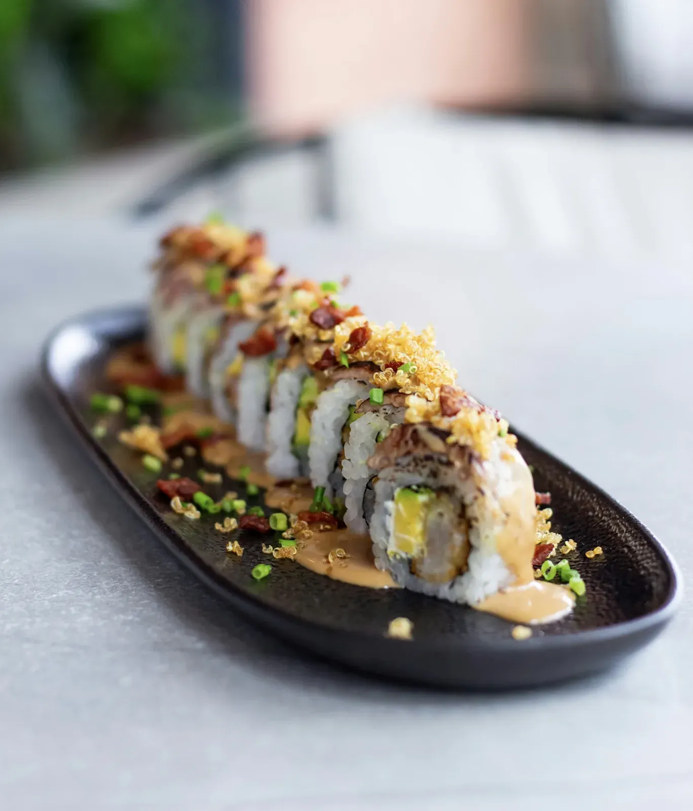 A long roll on a black plate, topped with torched eel, crunchy bits and scallion, shrimp tempura and avocado inside