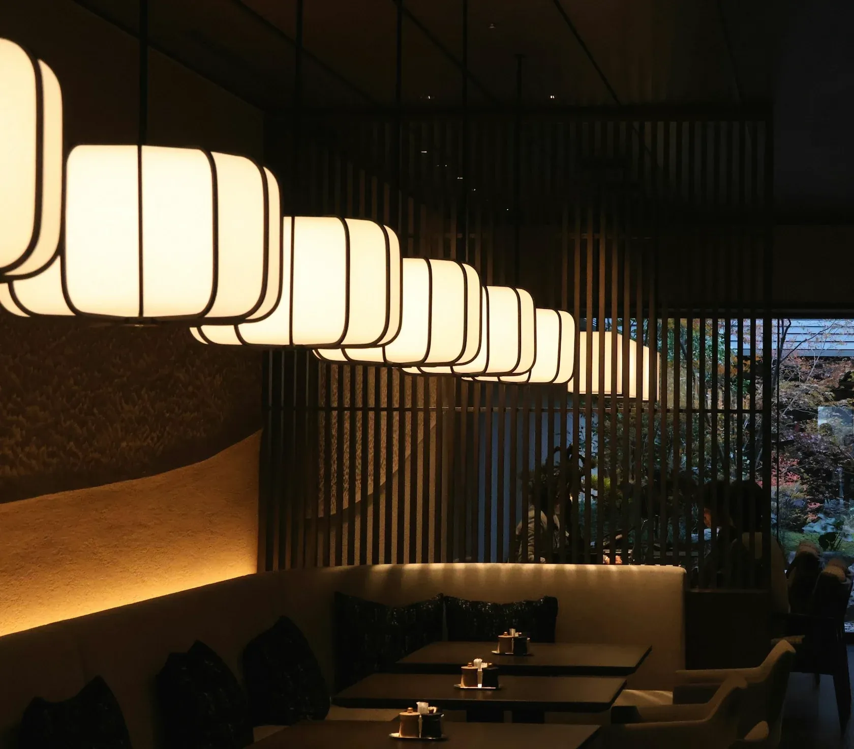 A dim dining room under a row of paper lanterns, with slatted screens and a garden window