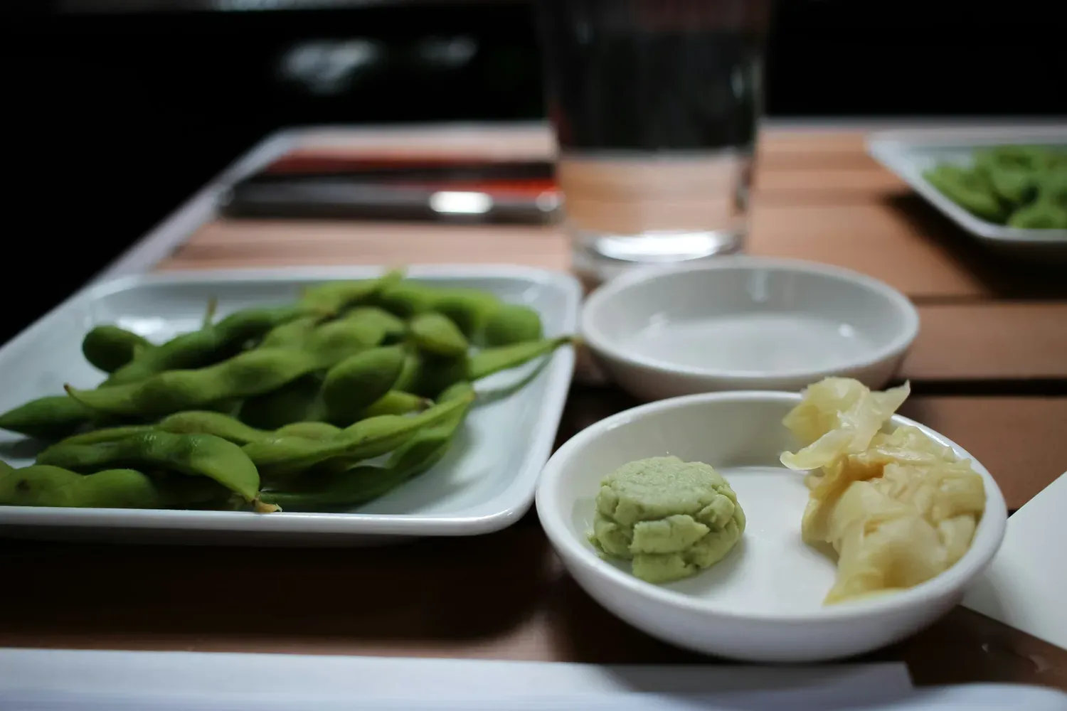 A plate of salted edamame on a wooden counter beside small dishes of wasabi and ginger