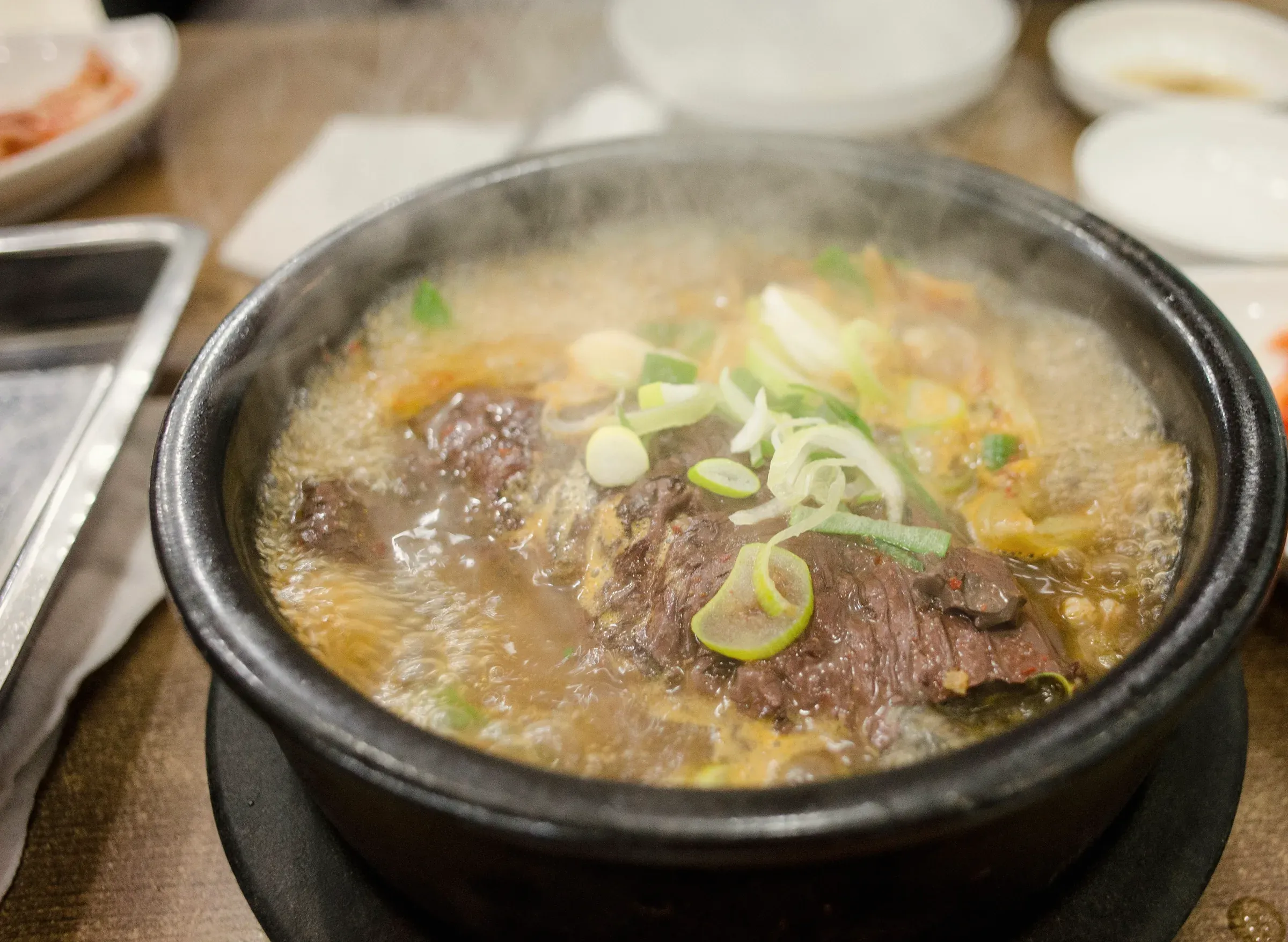 Short rib soup steaming in a black stone pot, with banchan behind