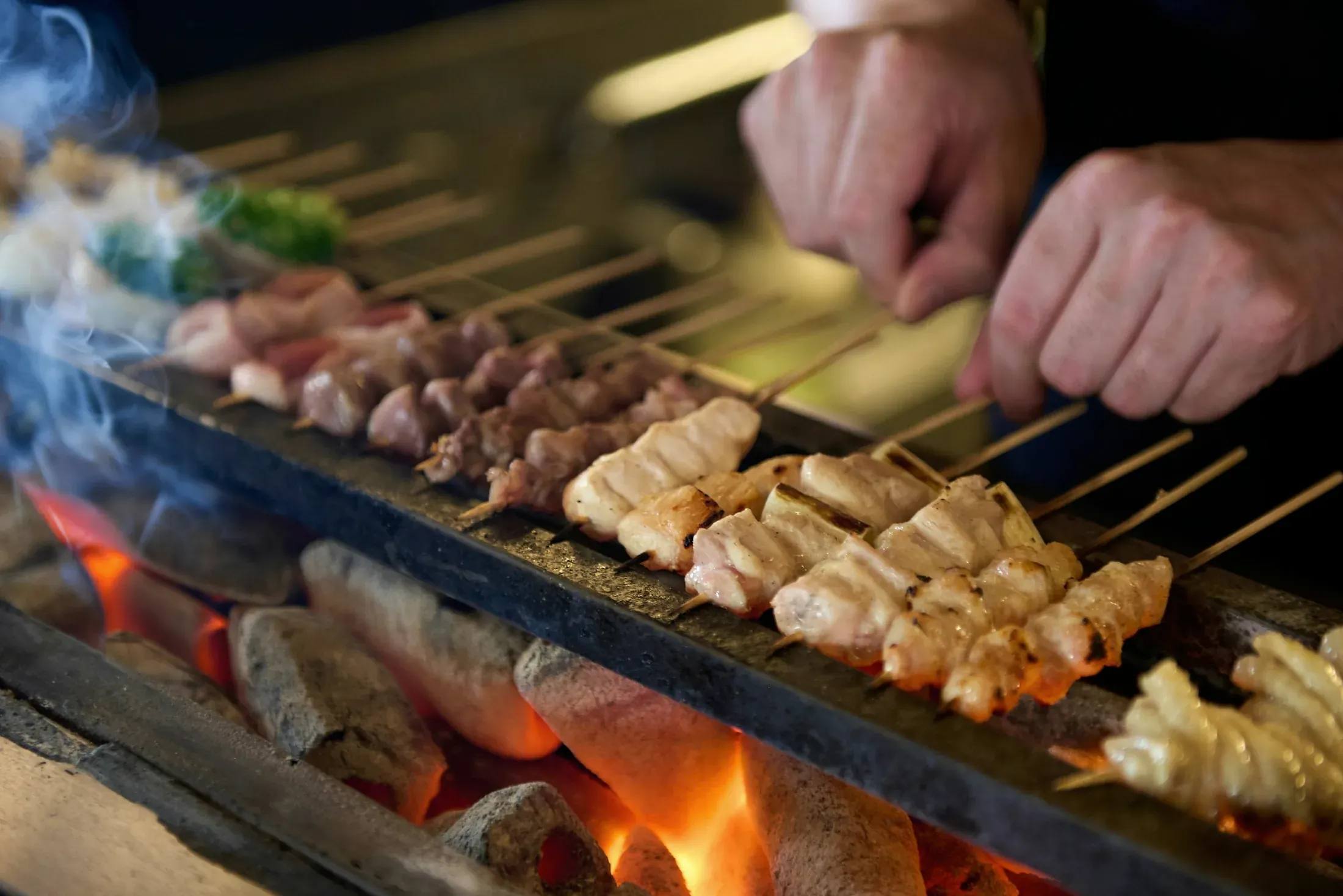 Chicken skewers over glowing white charcoal, the cook's hands laying another row on the grill