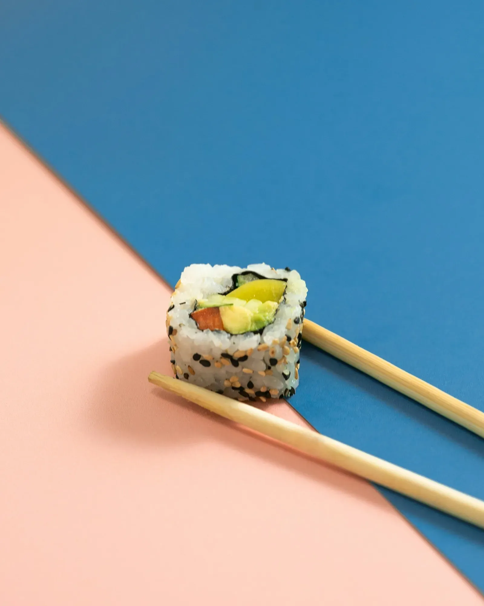One inside-out roll with avocado, salmon and cucumber, held by wooden chopsticks over a split blue and peach background