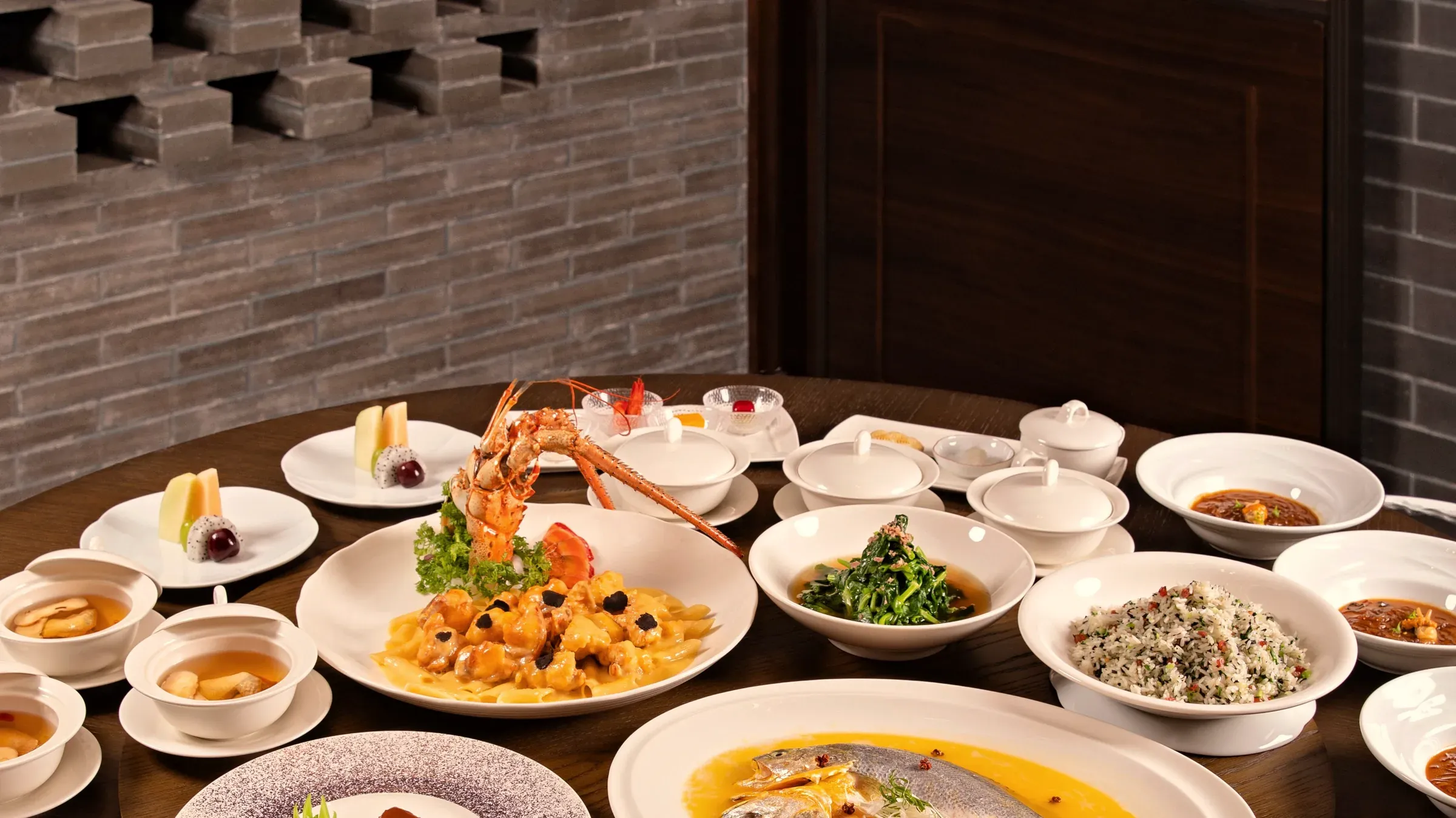A round banquet table set on a lazy susan before a brick lattice wall: lobster, steamed fish, soups and small plates