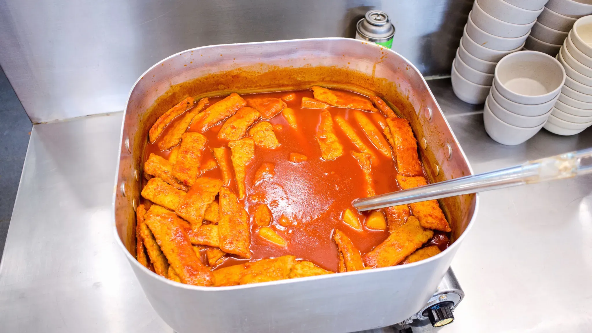 A steel pan of tteokbokki in red sauce on the shop counter, stacked bowls beside it