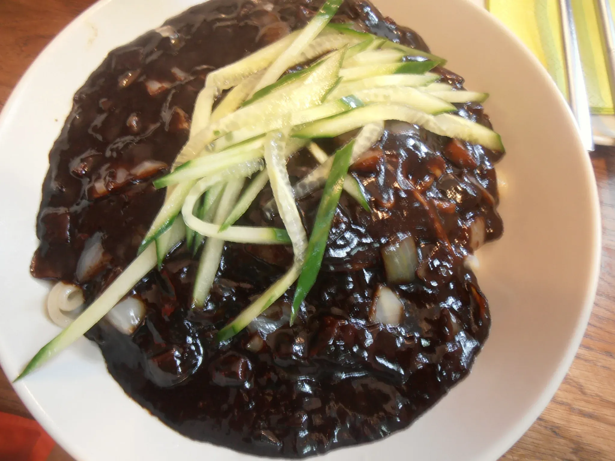 A bowl of jajangmyeon under glossy black bean sauce with diced onion and cucumber on top