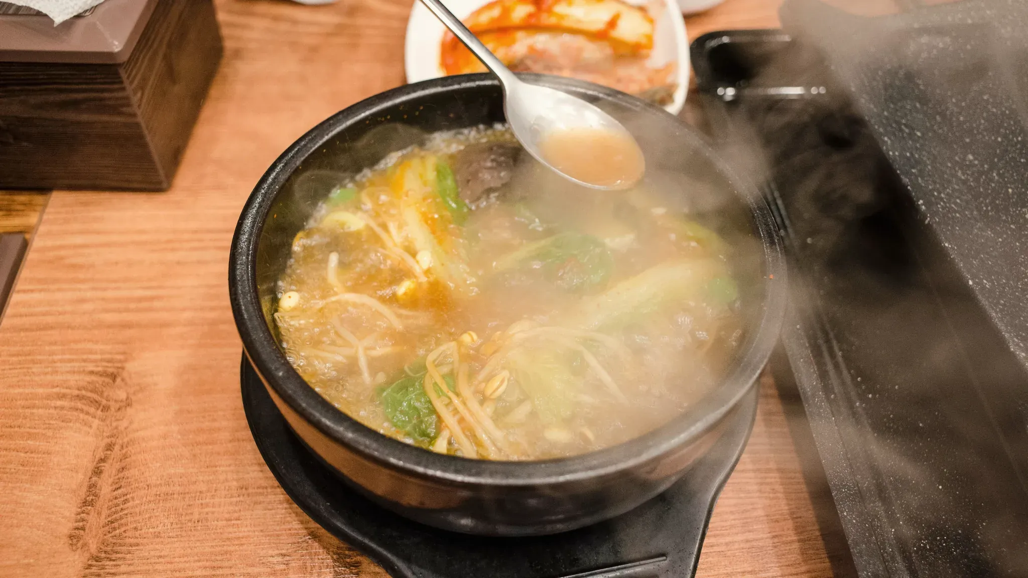 A ttukbaegi of gukbap steaming on the table, a spoon lifting the broth