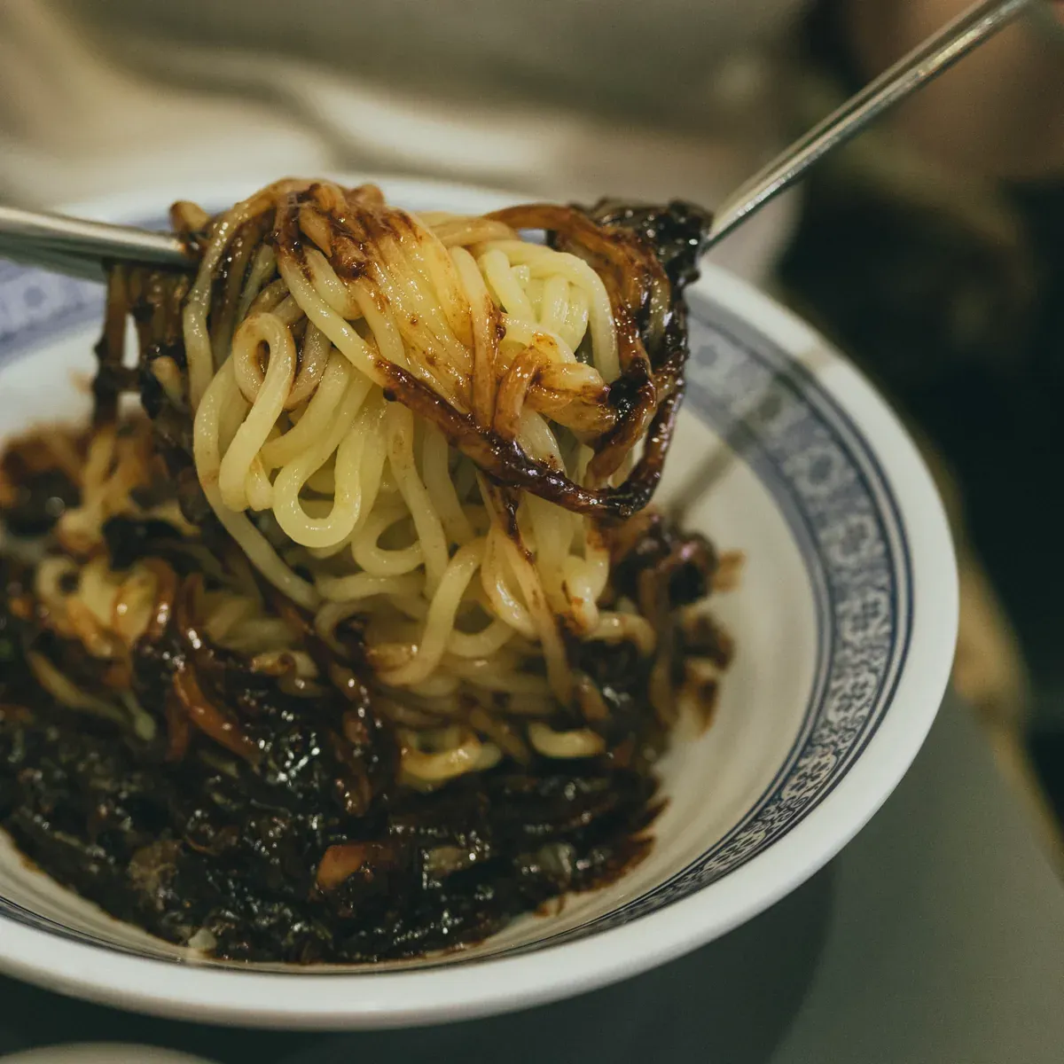 Noodles lifted out of a bowl of black bean sauce with chopsticks and a spoon