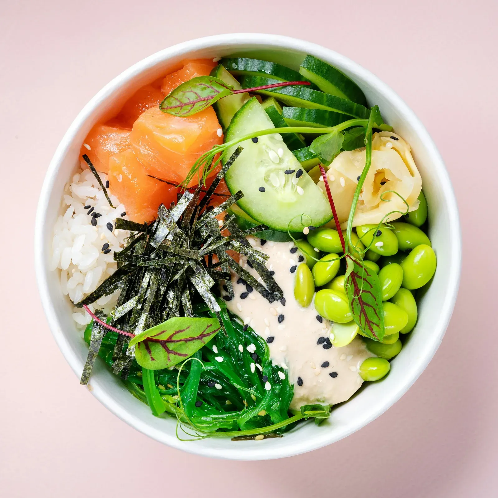 A poke bowl from above on pink: salmon, edamame, seaweed salad, cucumber, nori strips and sesame dressing