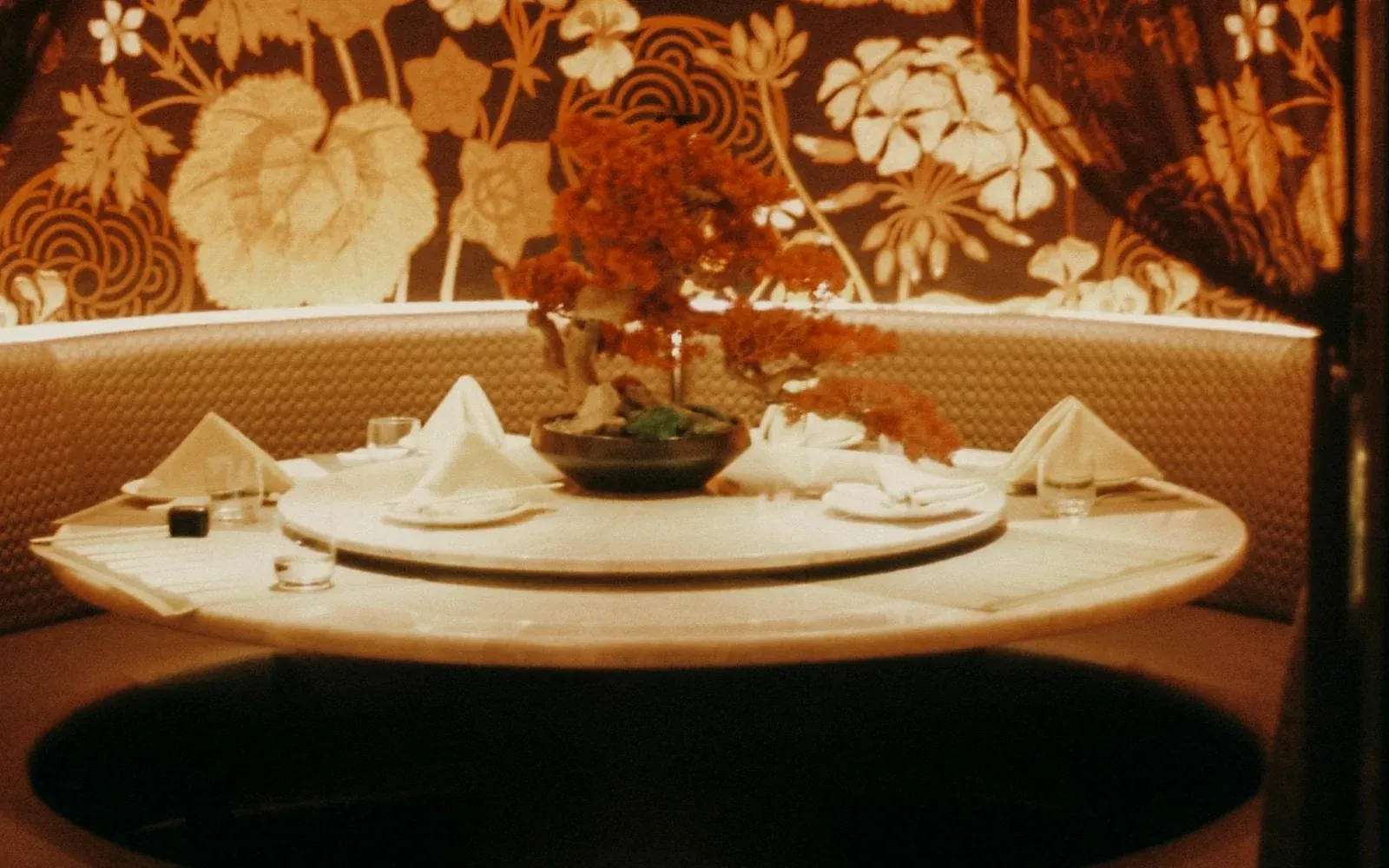 A curtained alcove with one round table set with folded napkins, a lazy susan and a bowl of red maple
