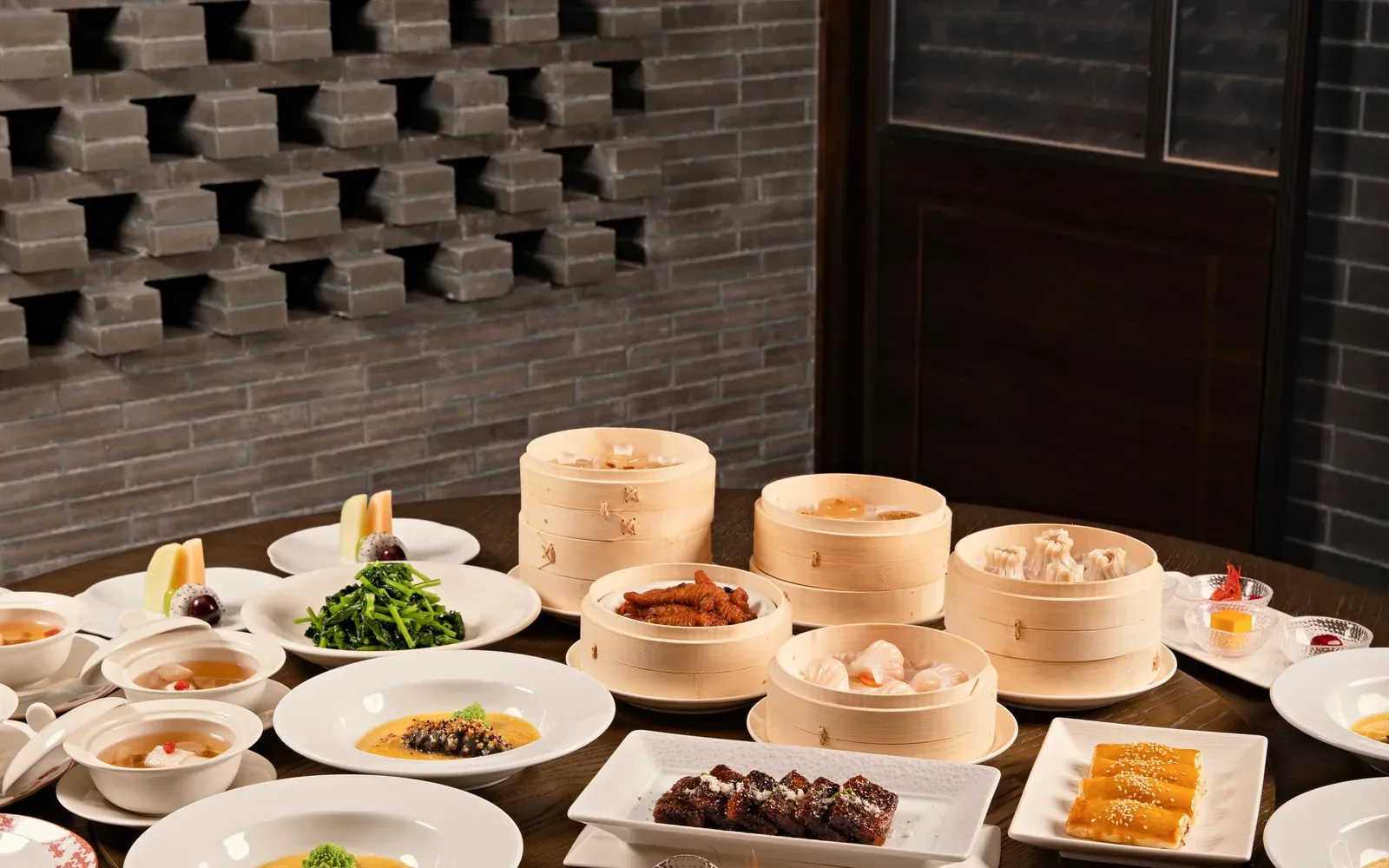 A large round table spread for a celebration with dim sum, soups and small plates, before a brick lattice wall