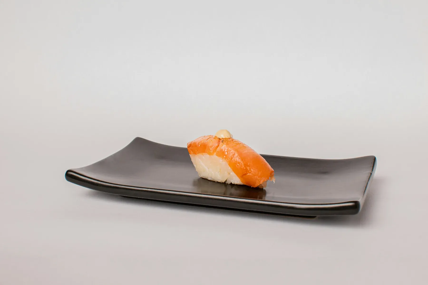 A single piece of salmon nigiri on a long black plate against a white wall