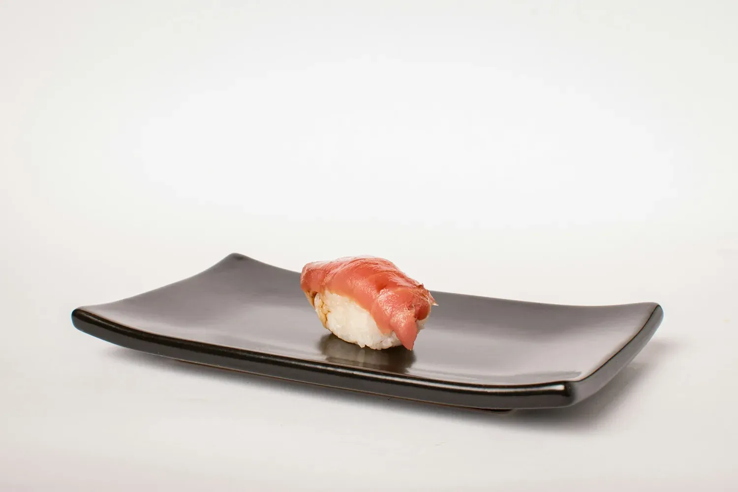A single piece of tuna nigiri on a long black plate against a white wall