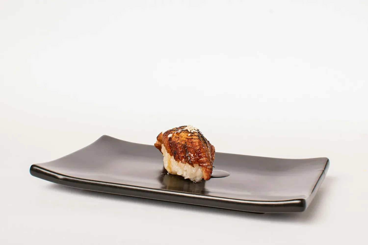 A single piece of glazed eel nigiri on a long black plate against a white wall