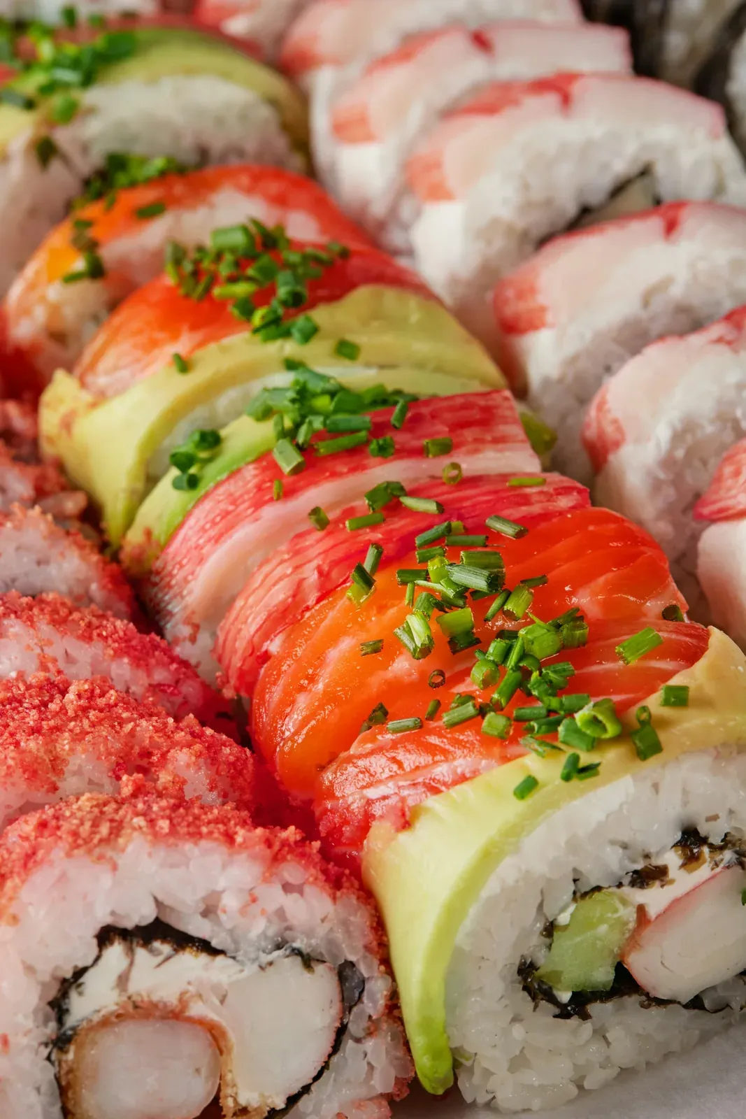 A long tray of rolls: red crunch rolls, rolls topped with salmon and avocado, and crab-topped rolls with chives