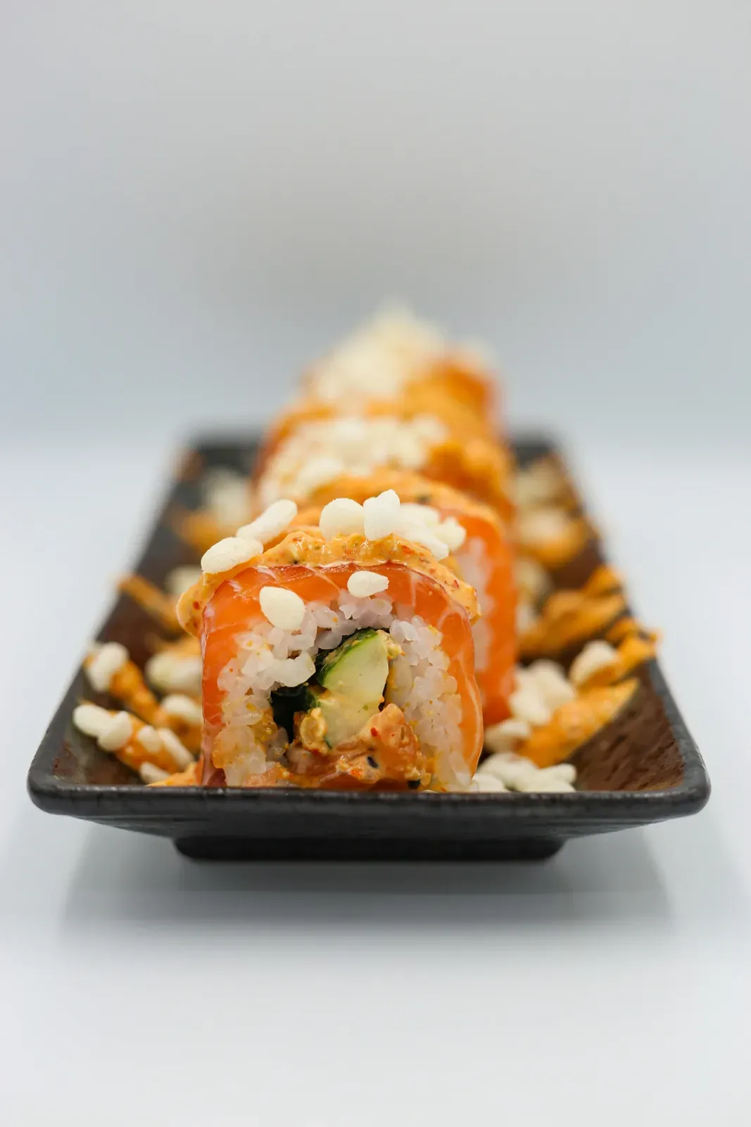 A salmon-topped roll filled with avocado and spicy salmon on a long dark plate, puffed rice and spicy mayo around it
