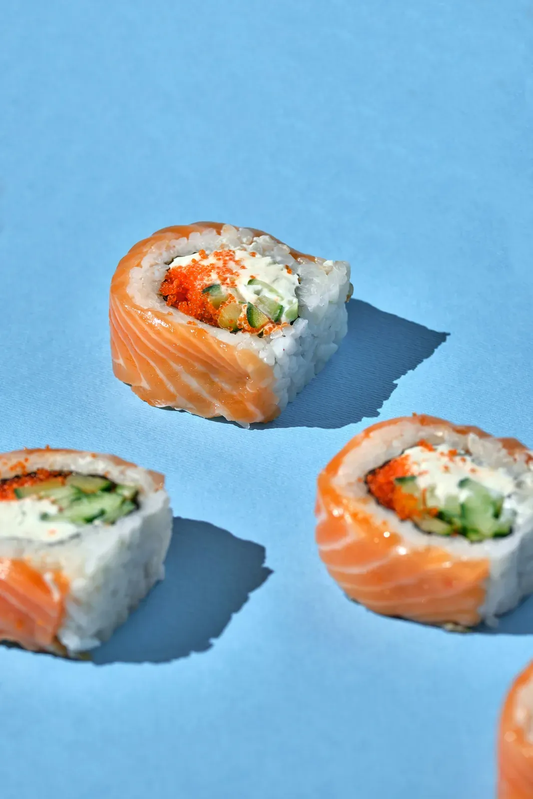 Three salmon-wrapped rolls on a sky blue table, filled with cream cheese, cucumber and orange masago