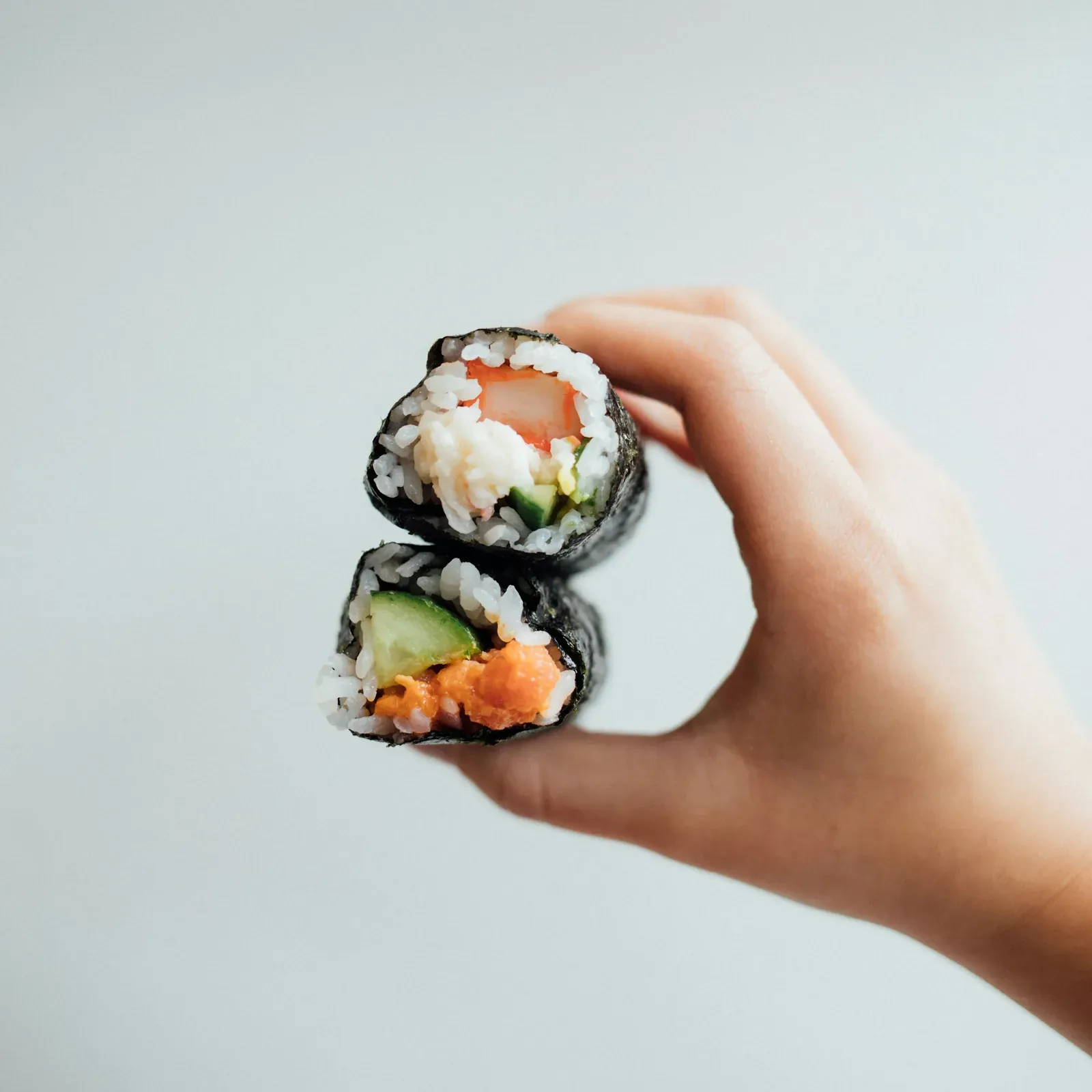 A hand holding two pieces of salmon and cucumber maki against a white wall