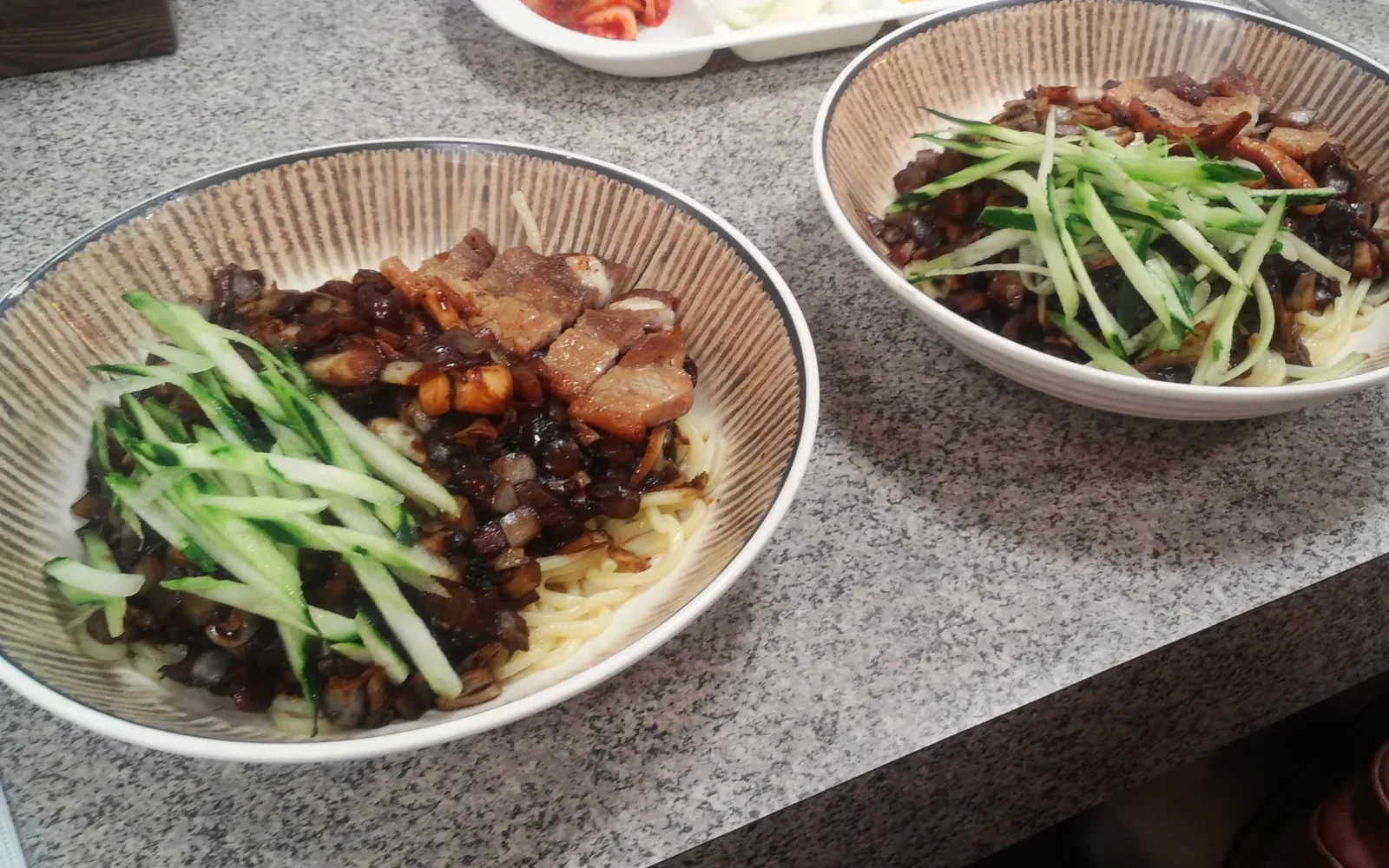 Two bowls of jajangmyeon with cucumber on a counter, kimchi, onion and danmuji behind