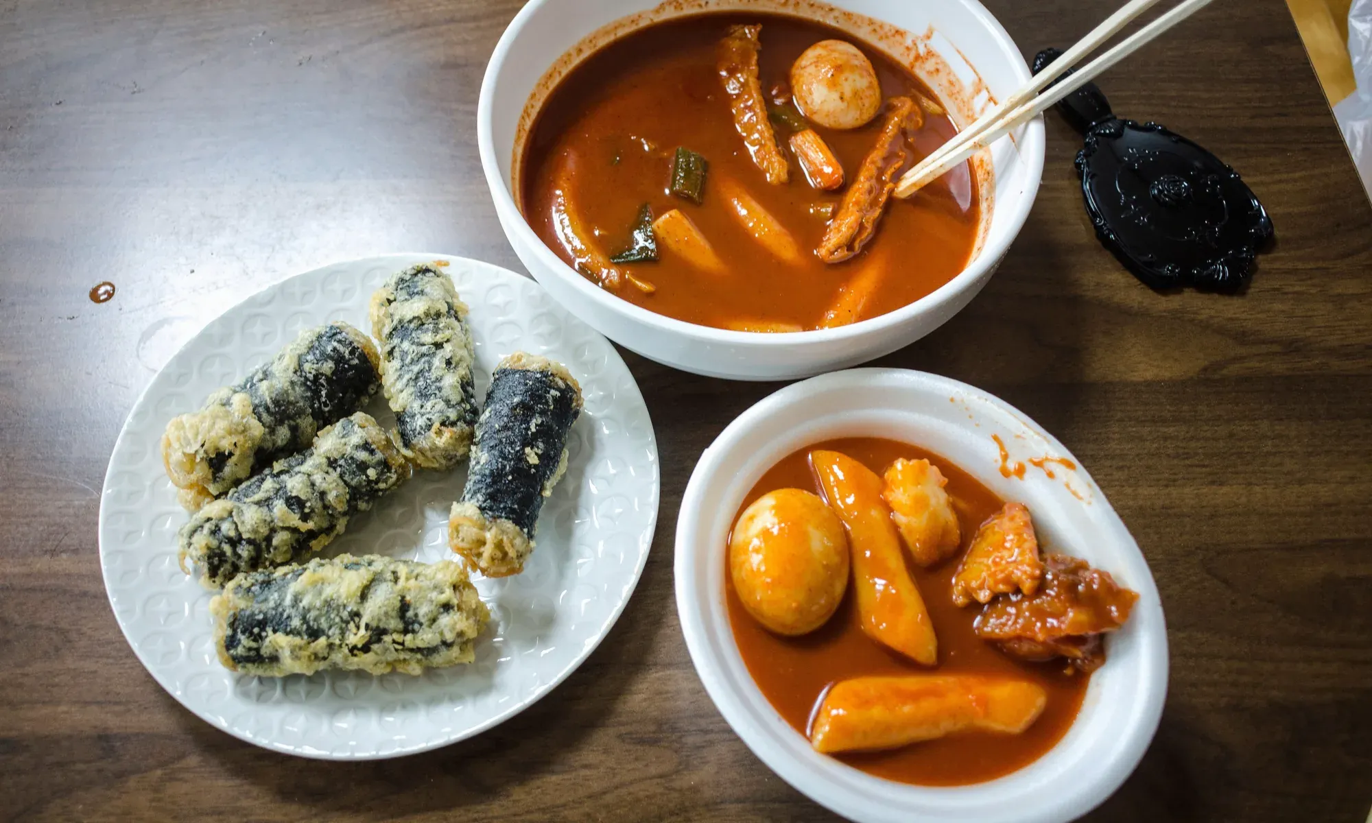 Two bowls of tteokbokki with boiled eggs and a plate of fried seaweed rolls, seen from above