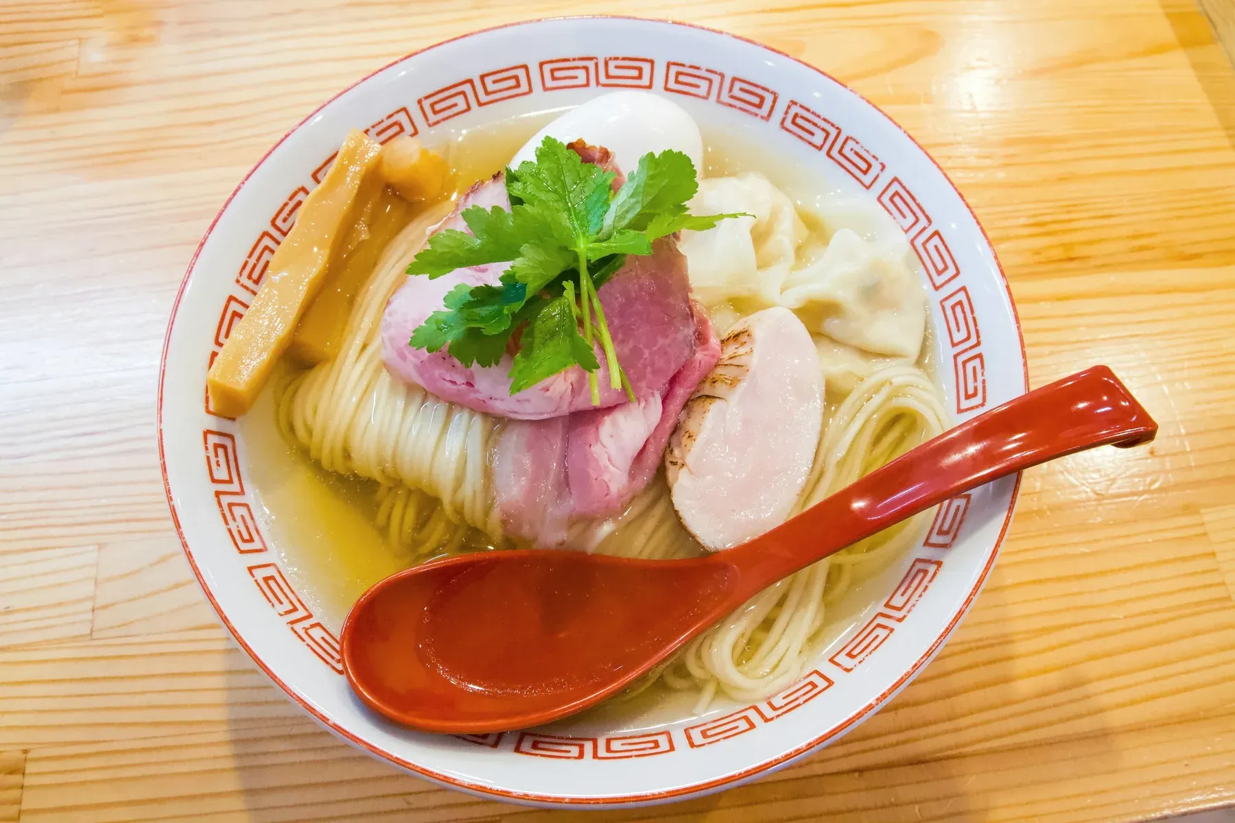 Clear shio ramen in a patterned bowl with fine noodles, pink pork and chicken chashu, a wonton, bamboo shoots and mitsuba, a red spoon resting in it