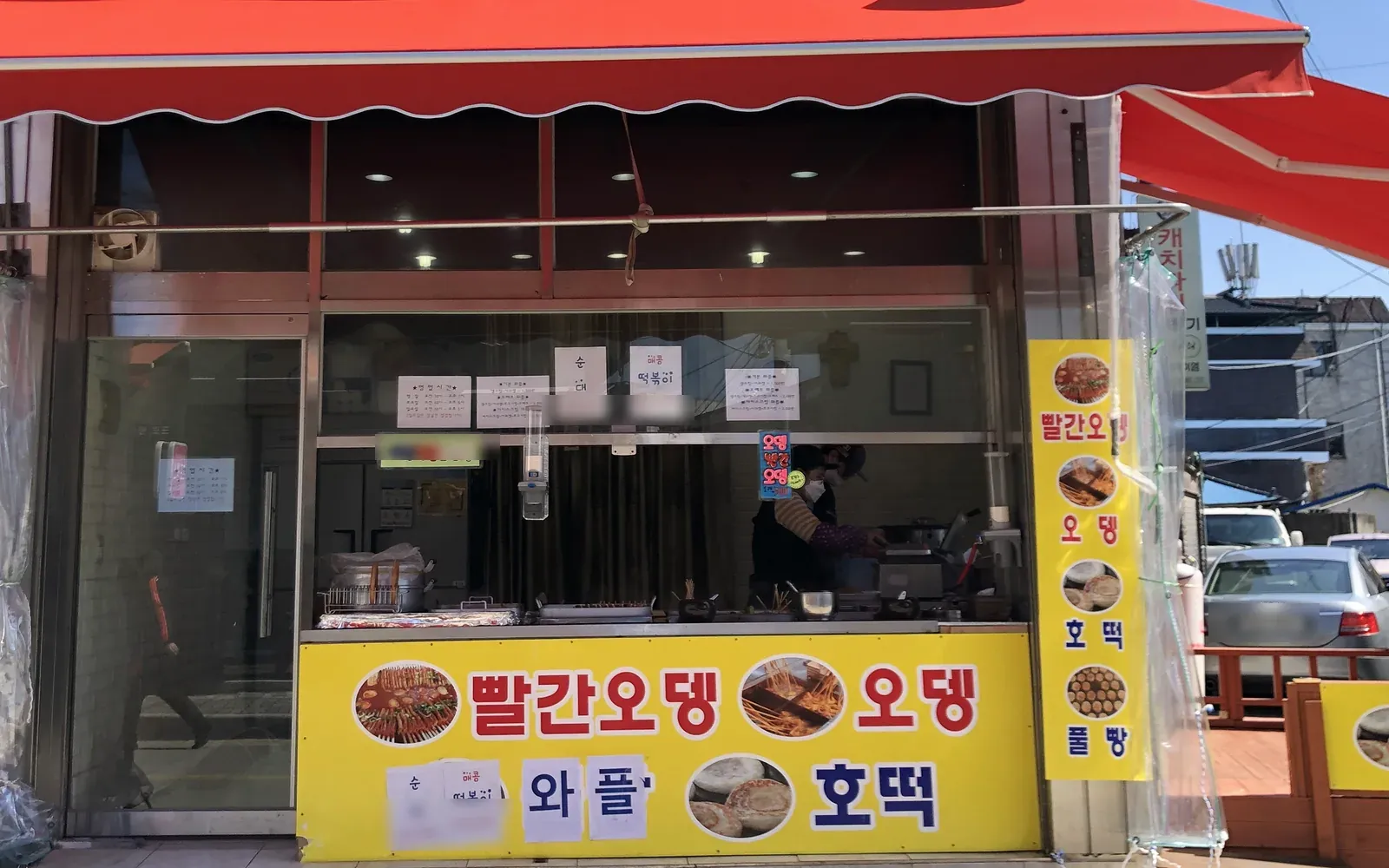 The takeout window under a red awning, fish cake and hotteok banners below the counter