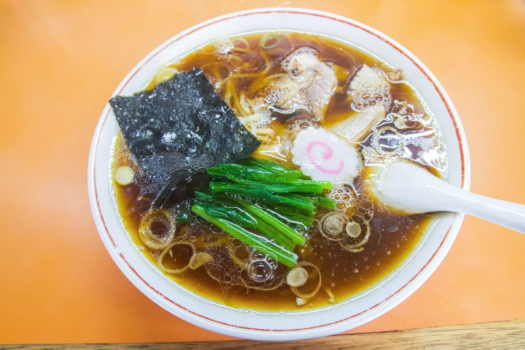 Dark amber shoyu ramen with a sheet of nori, a naruto slice, spinach, chashu and scallion rings