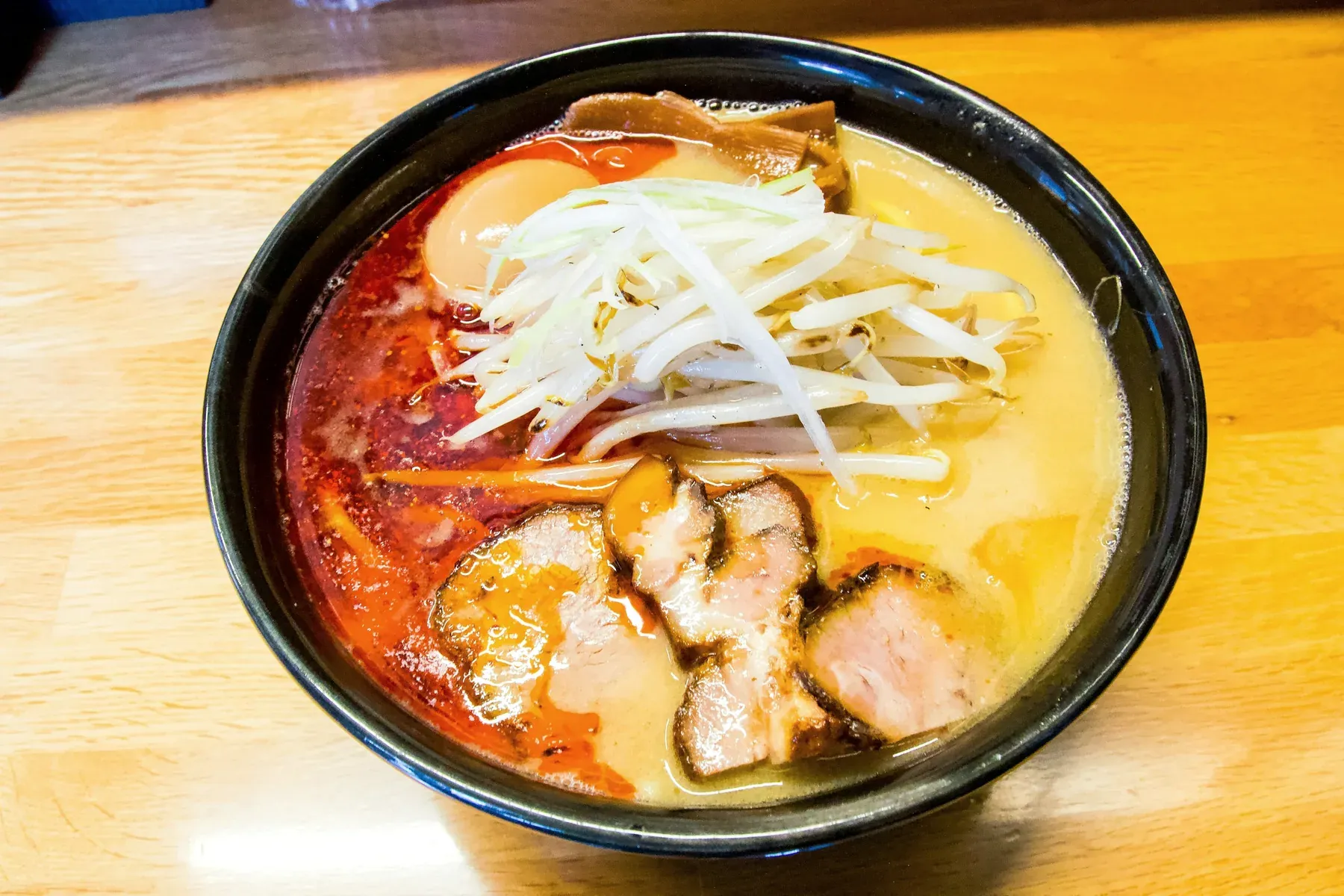 Spicy miso ramen in a black bowl, red chili oil on one side, bean sprouts, charred chashu and a soft egg