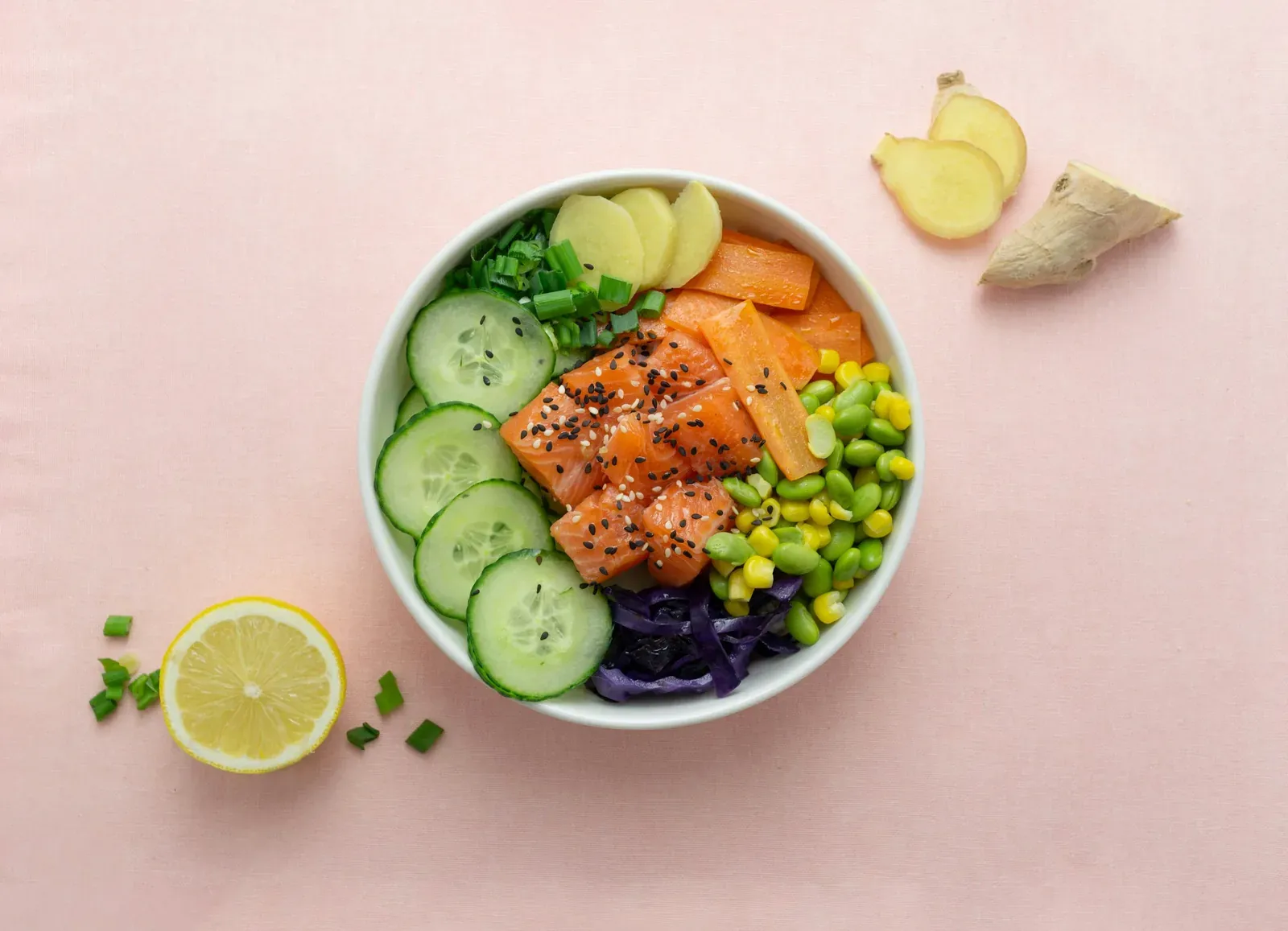 A spicy salmon poke bowl on a pink table with cucumber, corn, edamame and red cabbage, lemon and ginger beside it