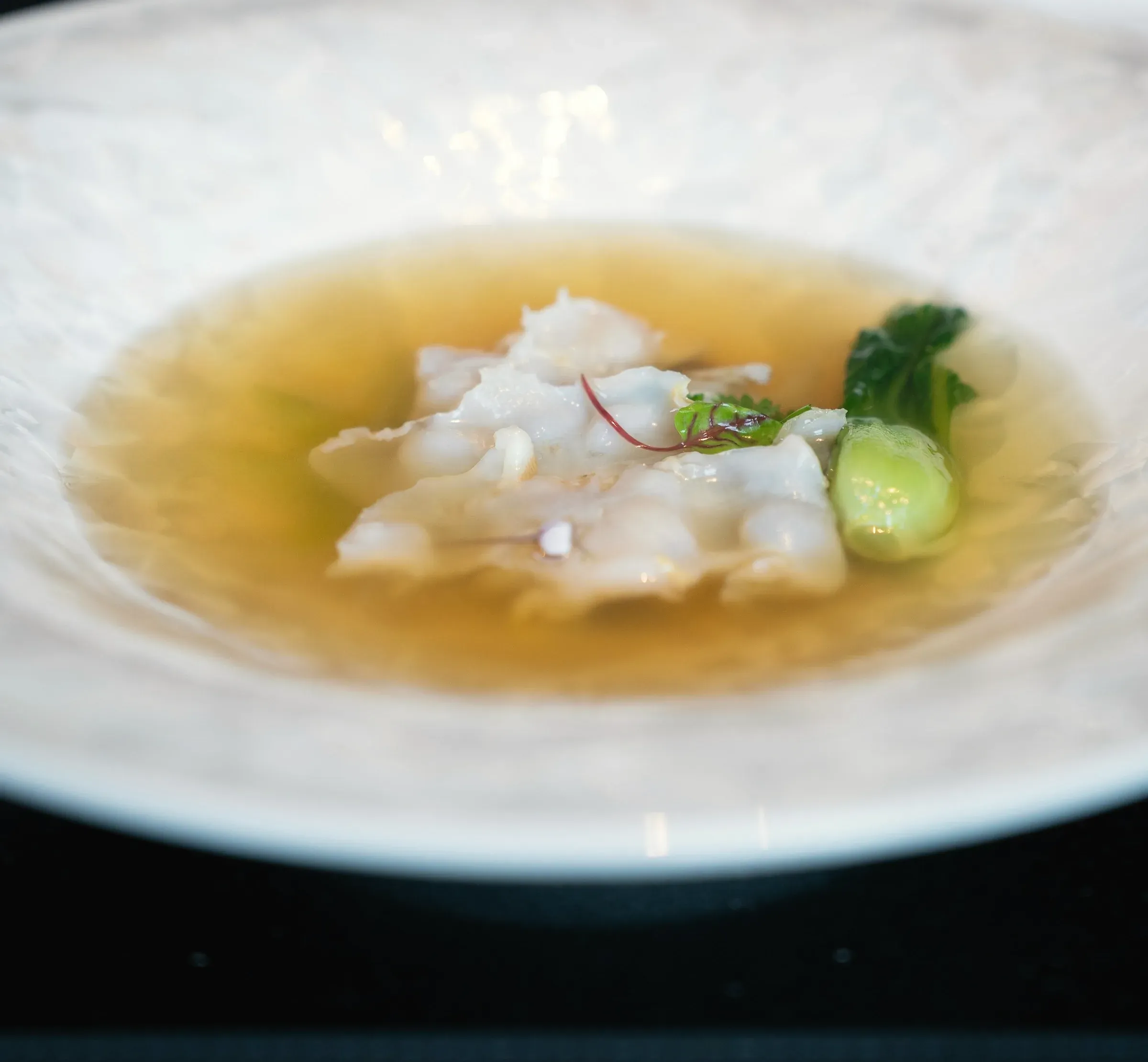 A slice of white sea bream and a sprig of greens in a clear golden broth, in a wide white bowl