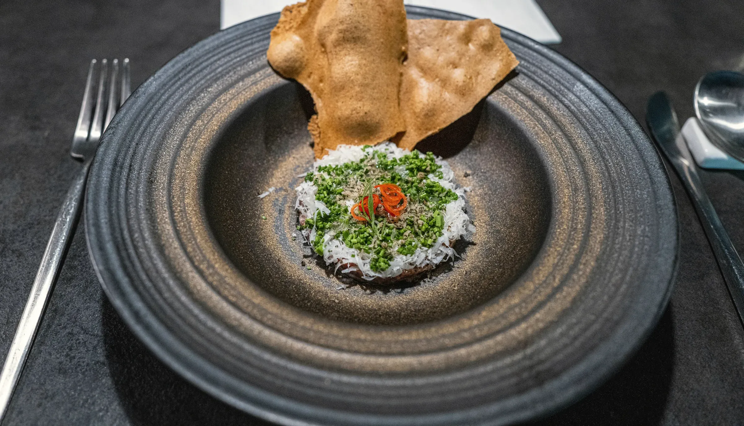Herb-topped soft tofu under two thin rice crisps on a dark stoneware plate