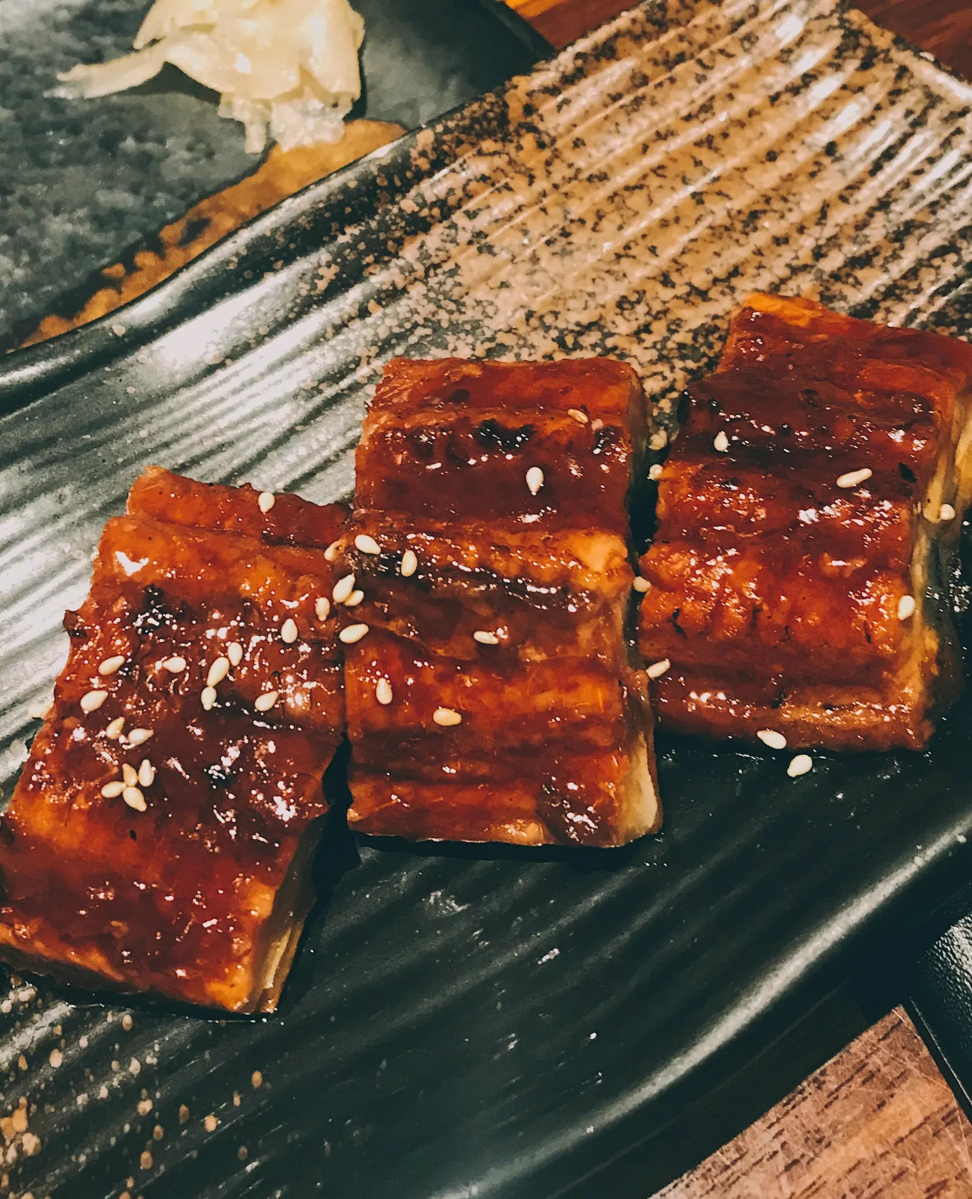 Three pieces of glazed grilled eel with sesame and pickled ginger on a ridged black plate