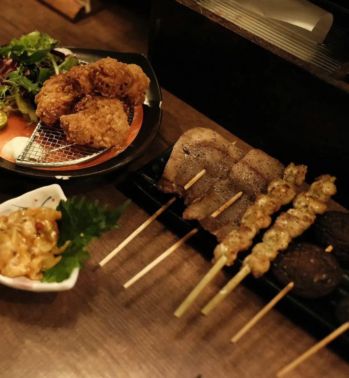 A table after work: skewers on a long plate, karaage on a wire rack, greens and a small dish of potato salad