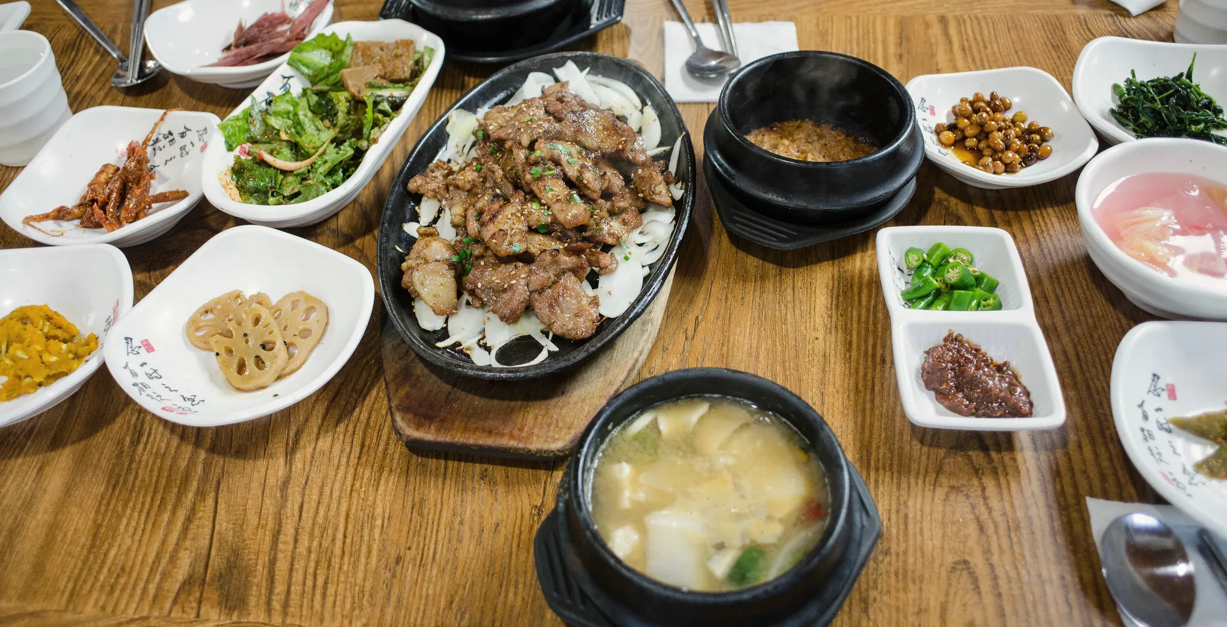 A long table set with banchan, stone pots of stew and a sizzling platter