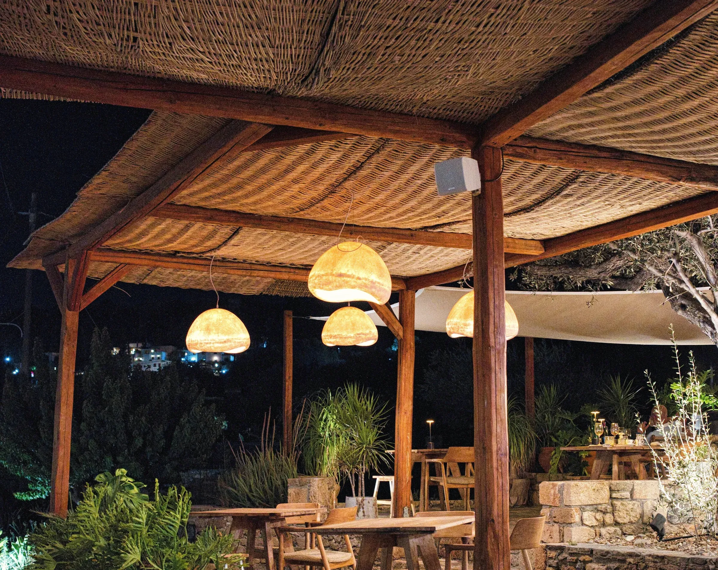 The terrace at night: oak tables on a stone floor under a reed-roofed pergola and soft pendant lamps