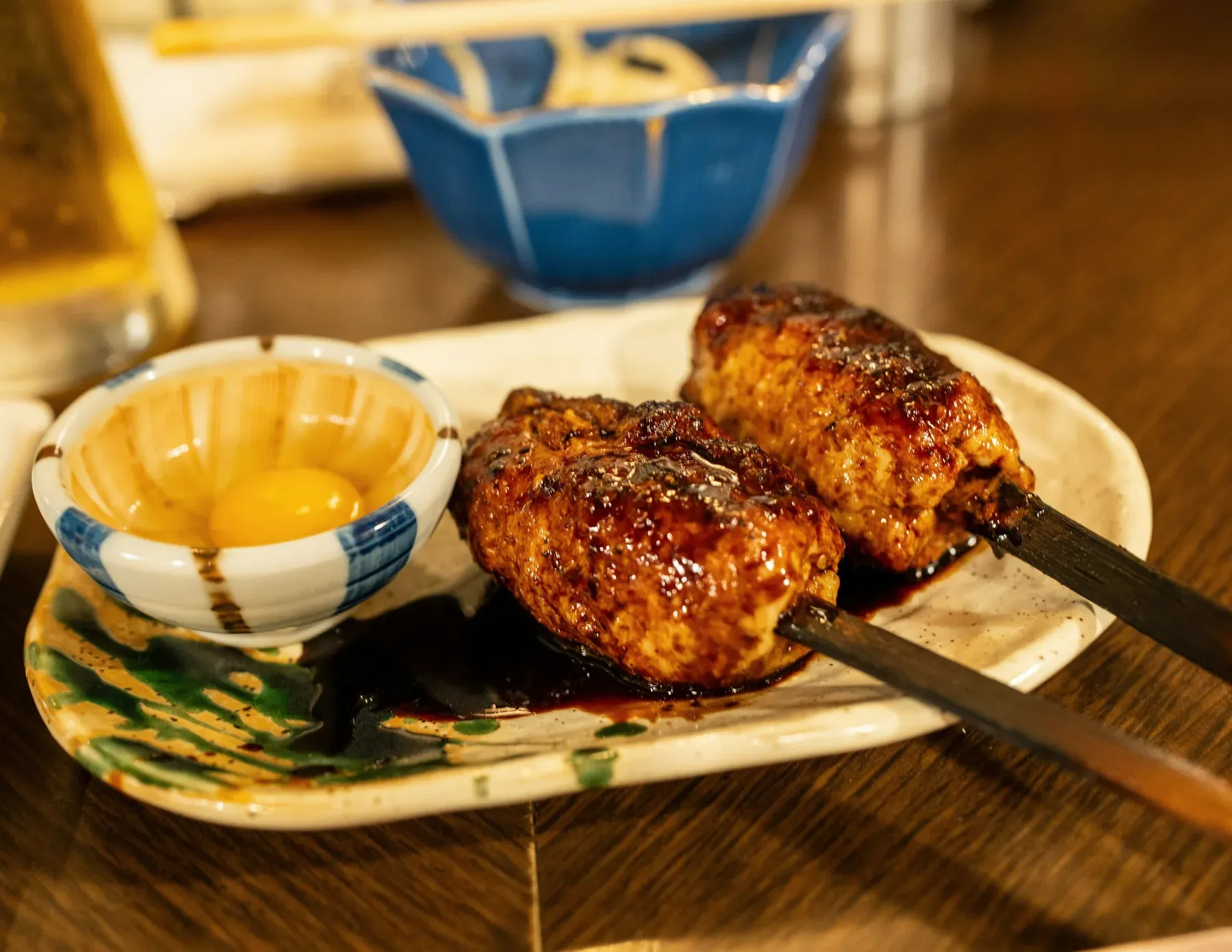 Two tsukune skewers glazed in tare beside a small bowl with a raw egg yolk