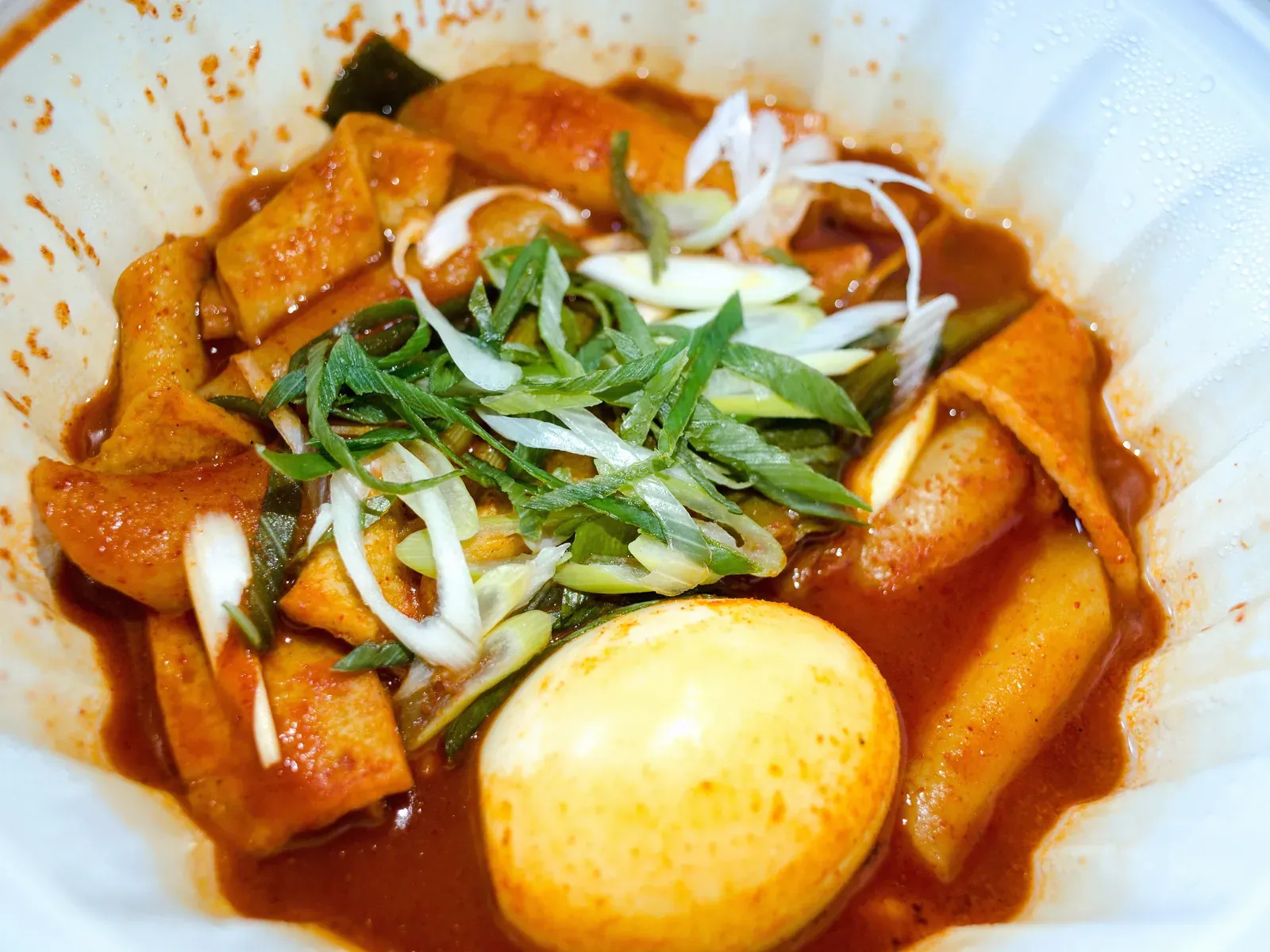 Rice cakes and fish cake in red sauce with scallion and a boiled egg in a takeout bowl