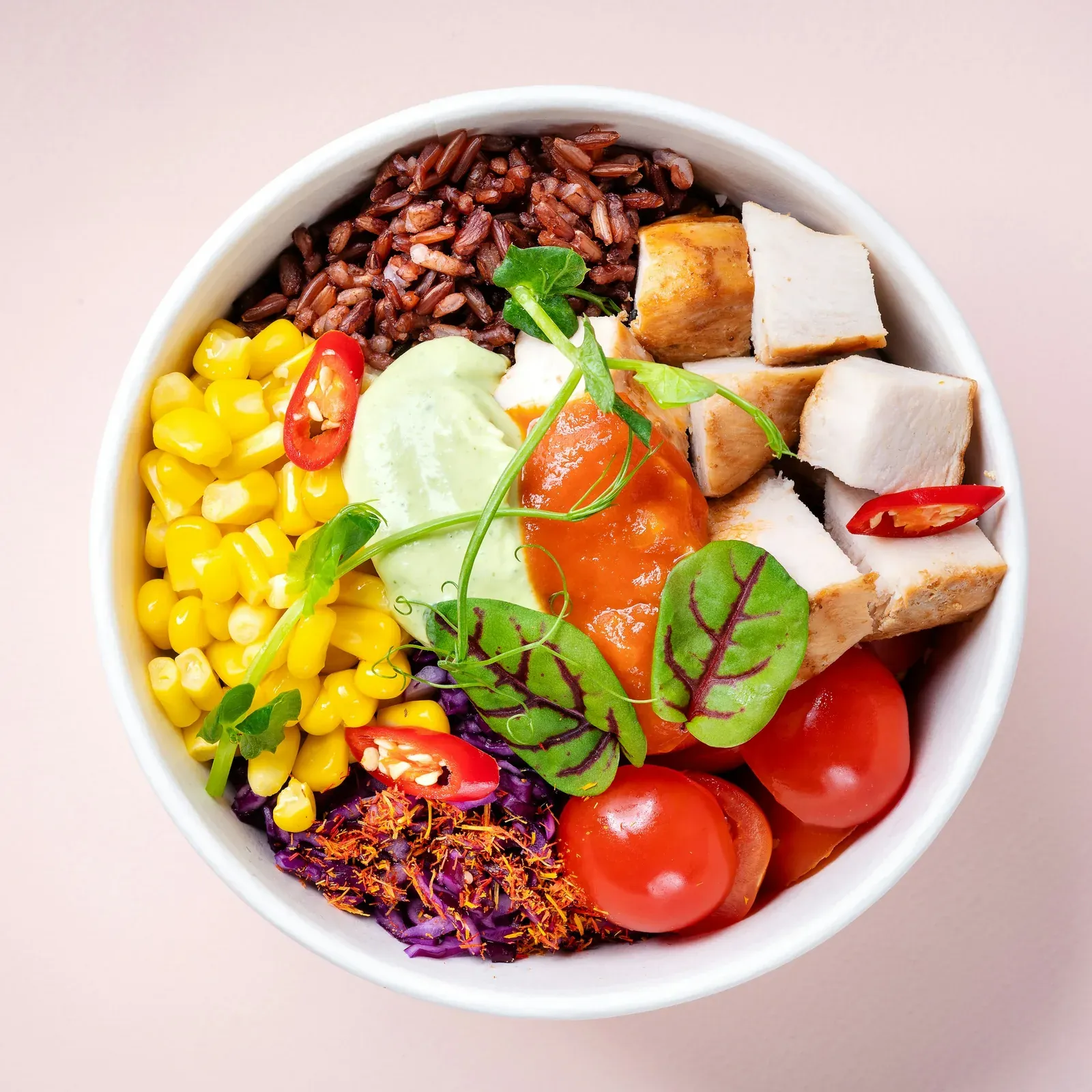 A poke bowl seen from above on a pink table: tofu, corn, cherry tomatoes, red cabbage and red chili over rice