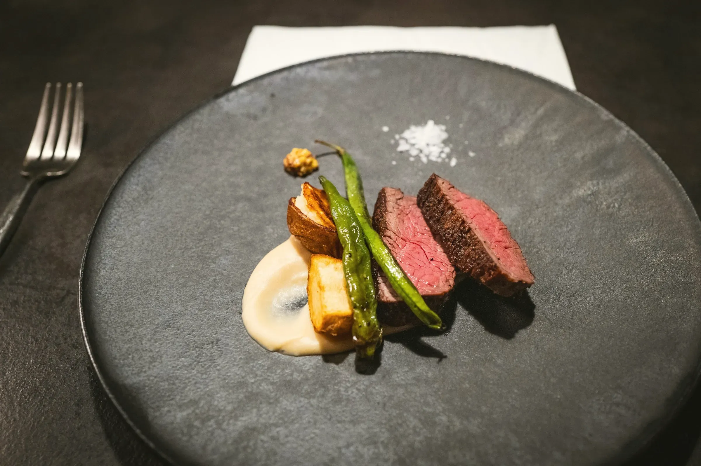 Charcoal-grilled beef striploin with grilled shishito, mushroom and a pale purée on a dark plate