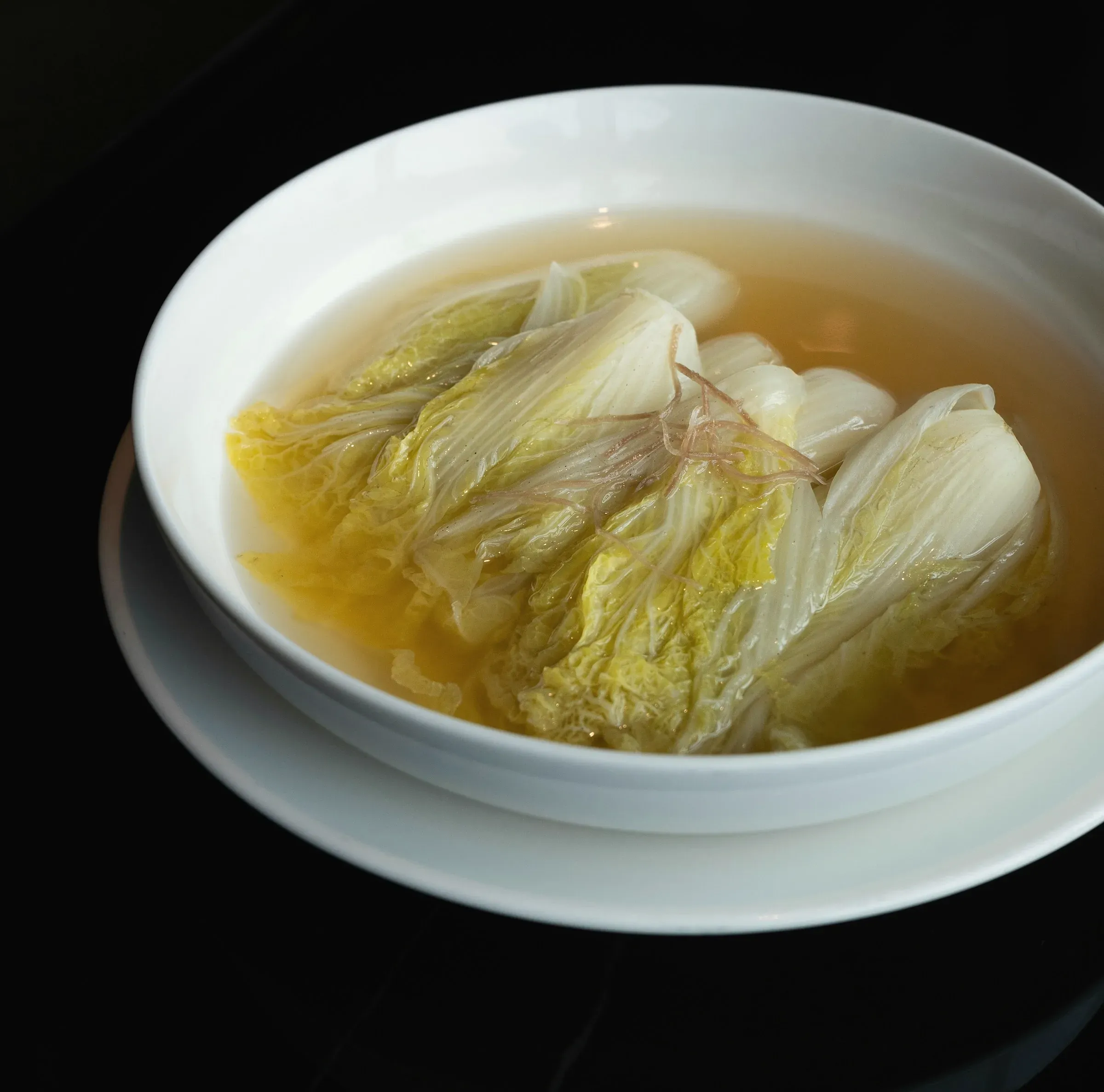 Soft napa cabbage leaves in a clear golden broth, in a white bowl on a saucer