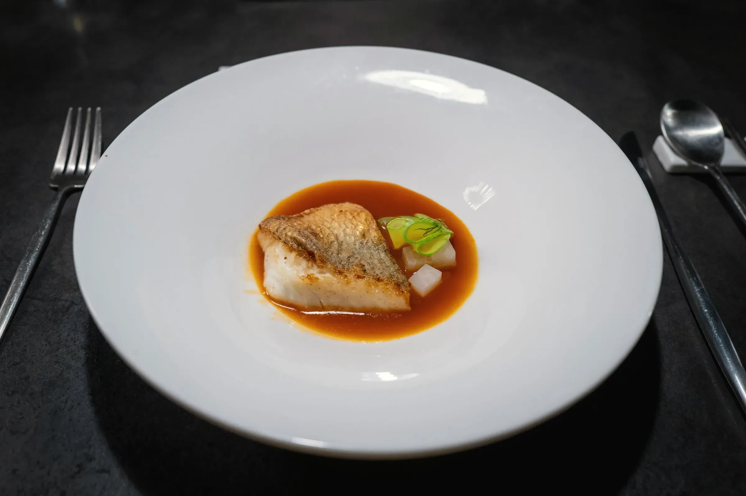 A seared cod fillet in a clear amber soy broth with cucumber and radish, in a wide white bowl