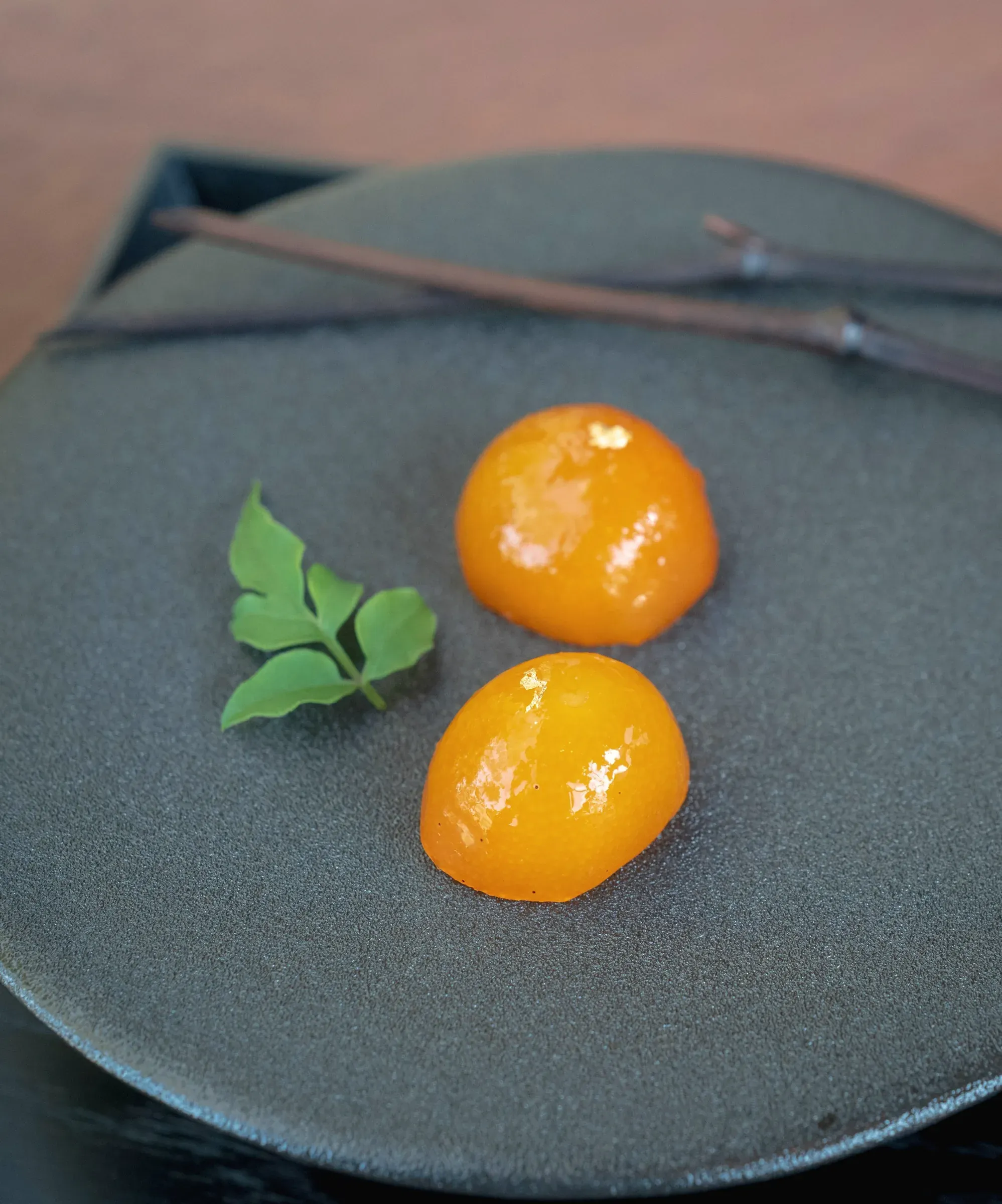 Two glossy candied kumquats and a sprig of leaves on dark stoneware